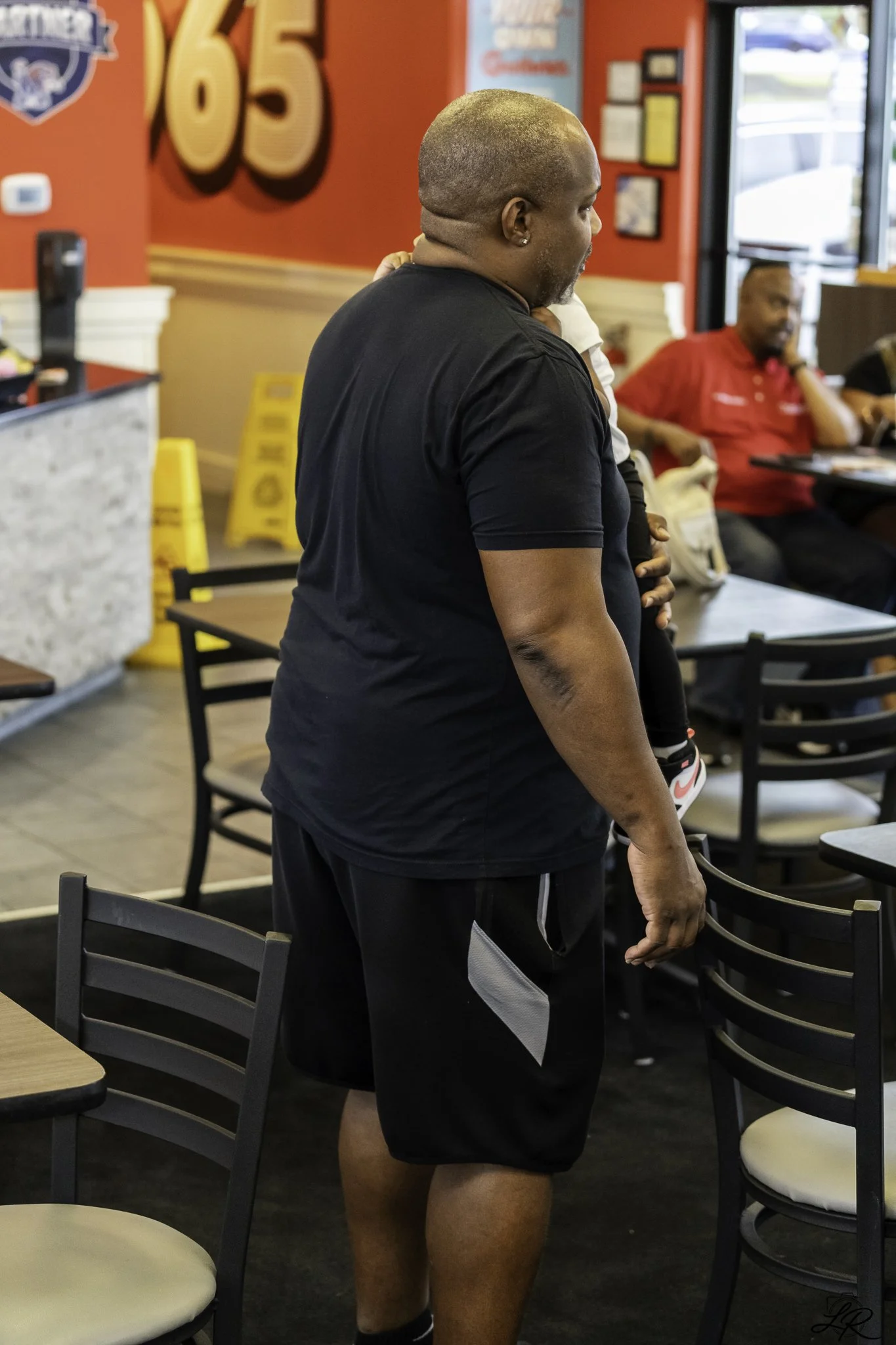A person with a shaved head and earrings is standing inside a restaurant, holding a baby dressed in black and white shoes. The background features red walls, yellow caution signs, and restaurant decor, with another man sitting in the background weari