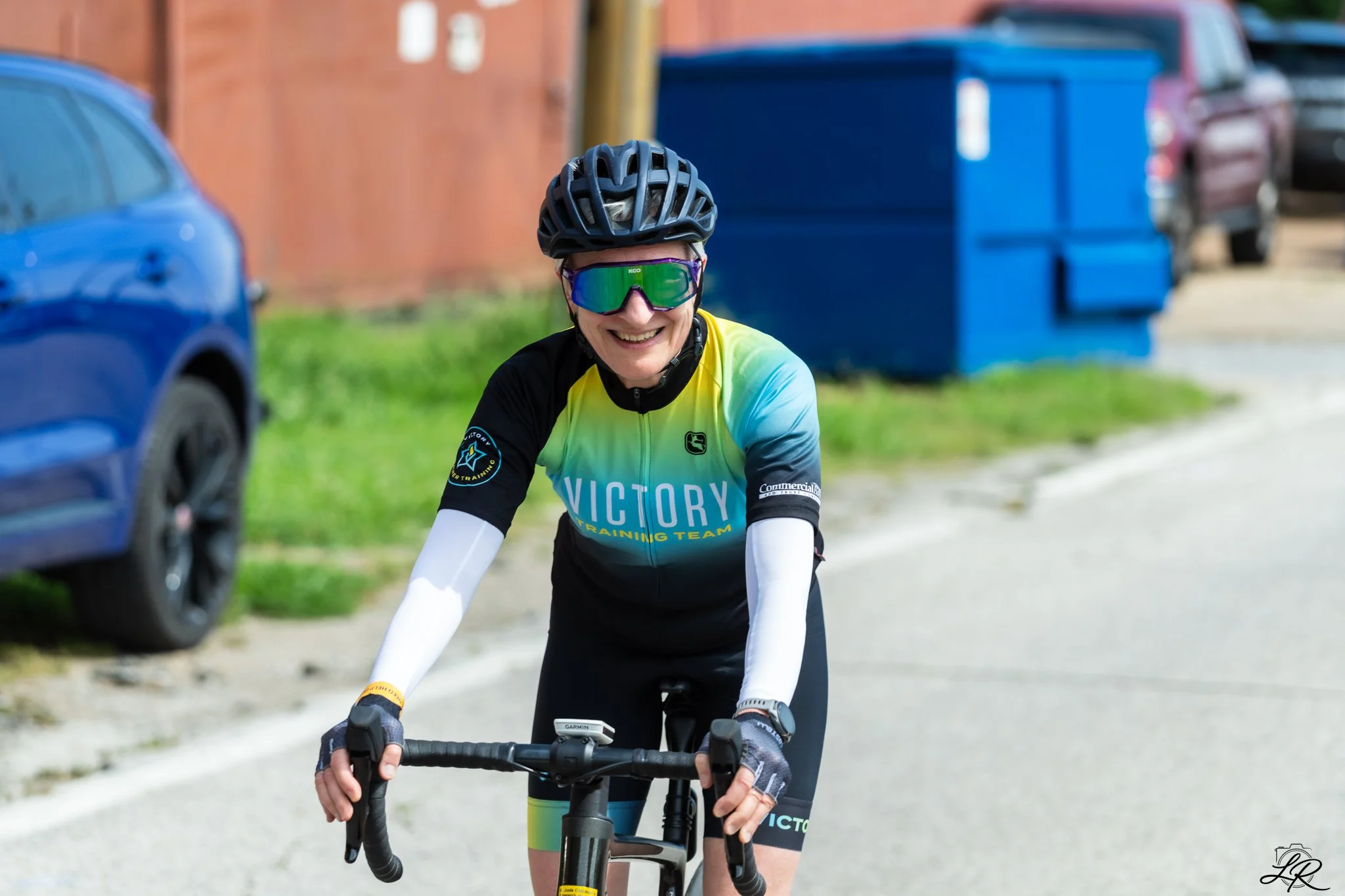A woman riding a bicycle outdoors, wearing a black helmet, colorful cycling jersey, sunglasses, and cycling gloves, with parked cars and a blue trash bin in the background.