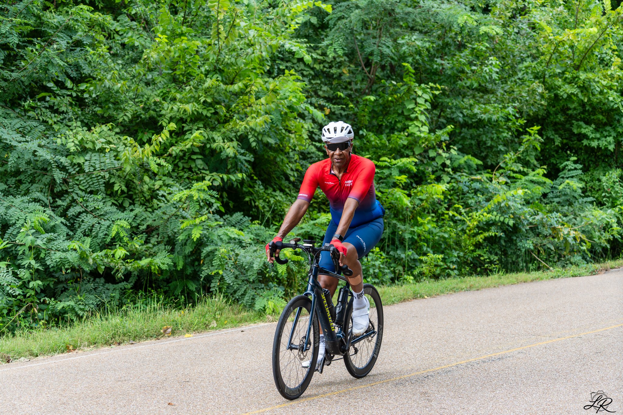 A man riding a black bicycle on a paved trail surrounded by lush green trees, wearing a red and blue cycling outfit, helmet, sunglasses, and white cycling shoes.