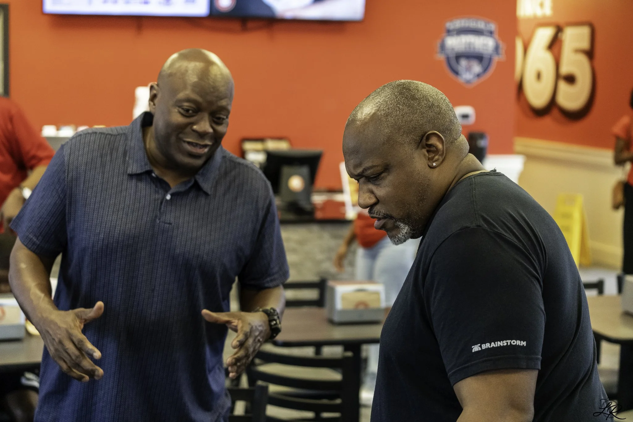 Two men engaging in conversation inside a fast-food restaurant with a red wall and a large '65' sign in the background.