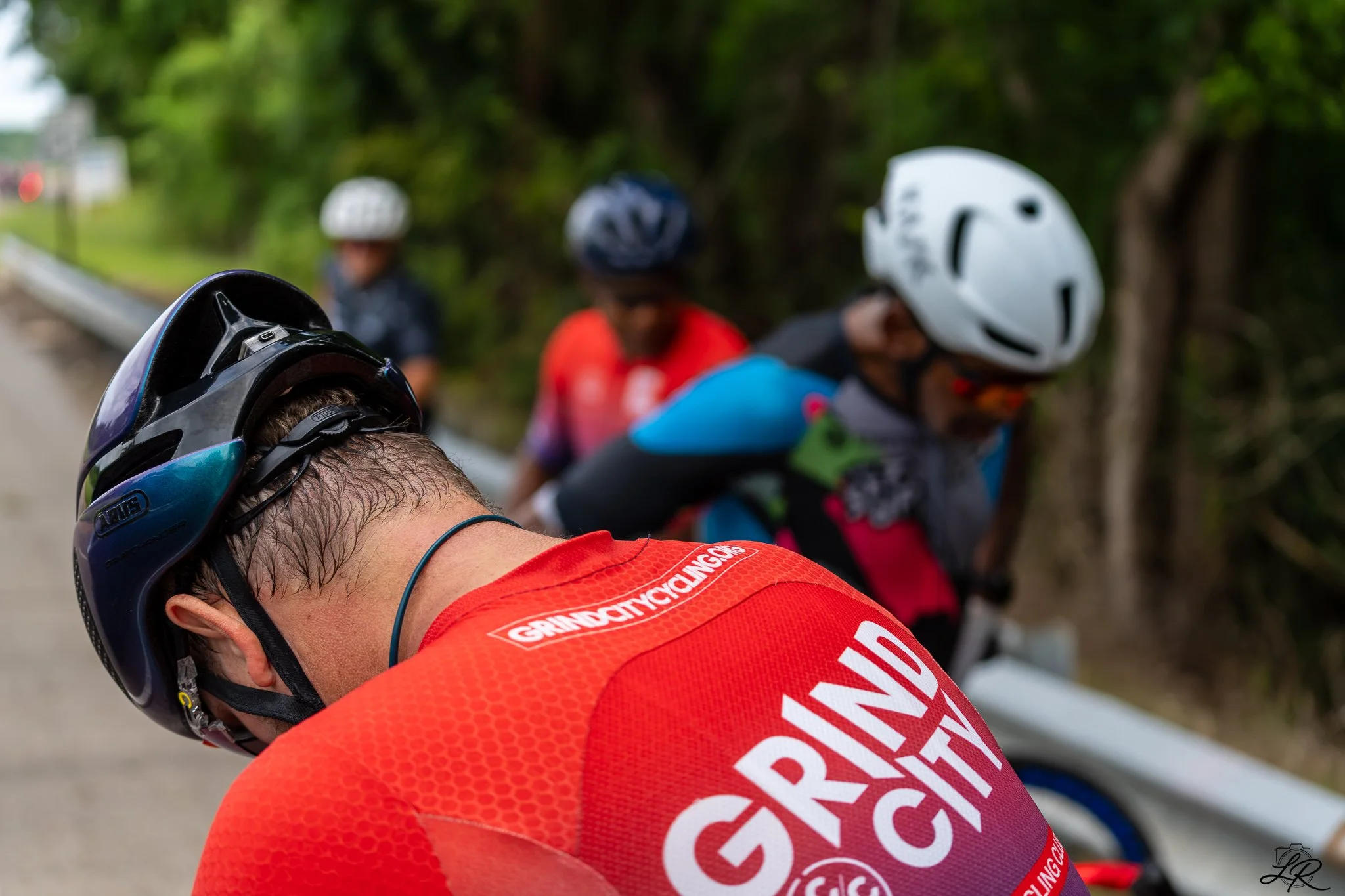Close-up of a man wearing a red cycling jersey and a black helmet, with other cyclists in helmets and colorful gear in the background, outdoors on a road with trees.