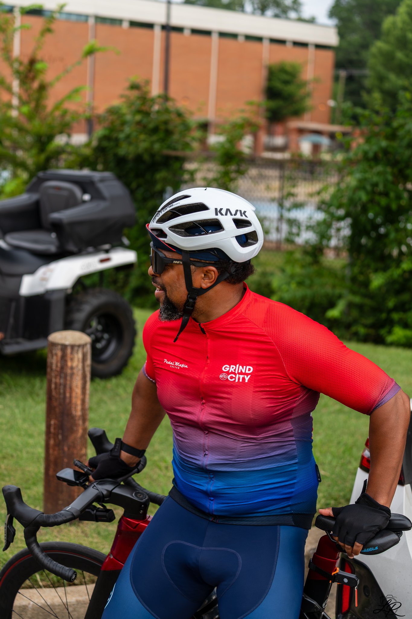 A man wearing a white bike helmet, red shirt, and blue cycling shorts standing with a bike outdoors near a parking lot with trees and a building in the background.