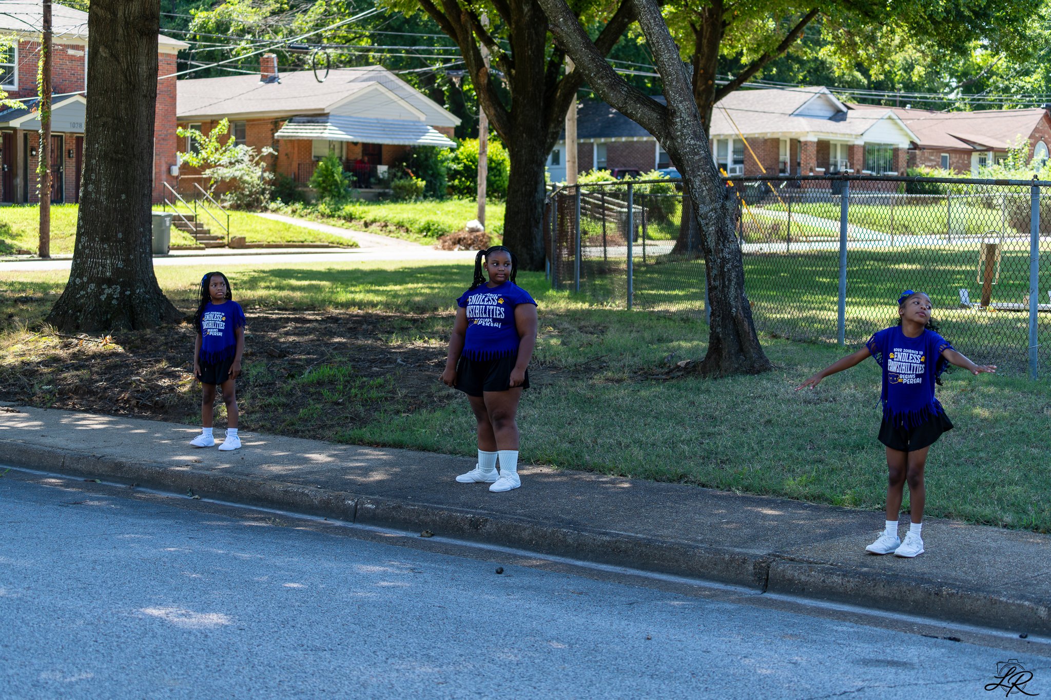 Three young girls wearing blue t-shirts and black shorts standing and stretching on a grassy area near a sidewalk, under large trees in a residential neighborhood with brick houses in the background.
