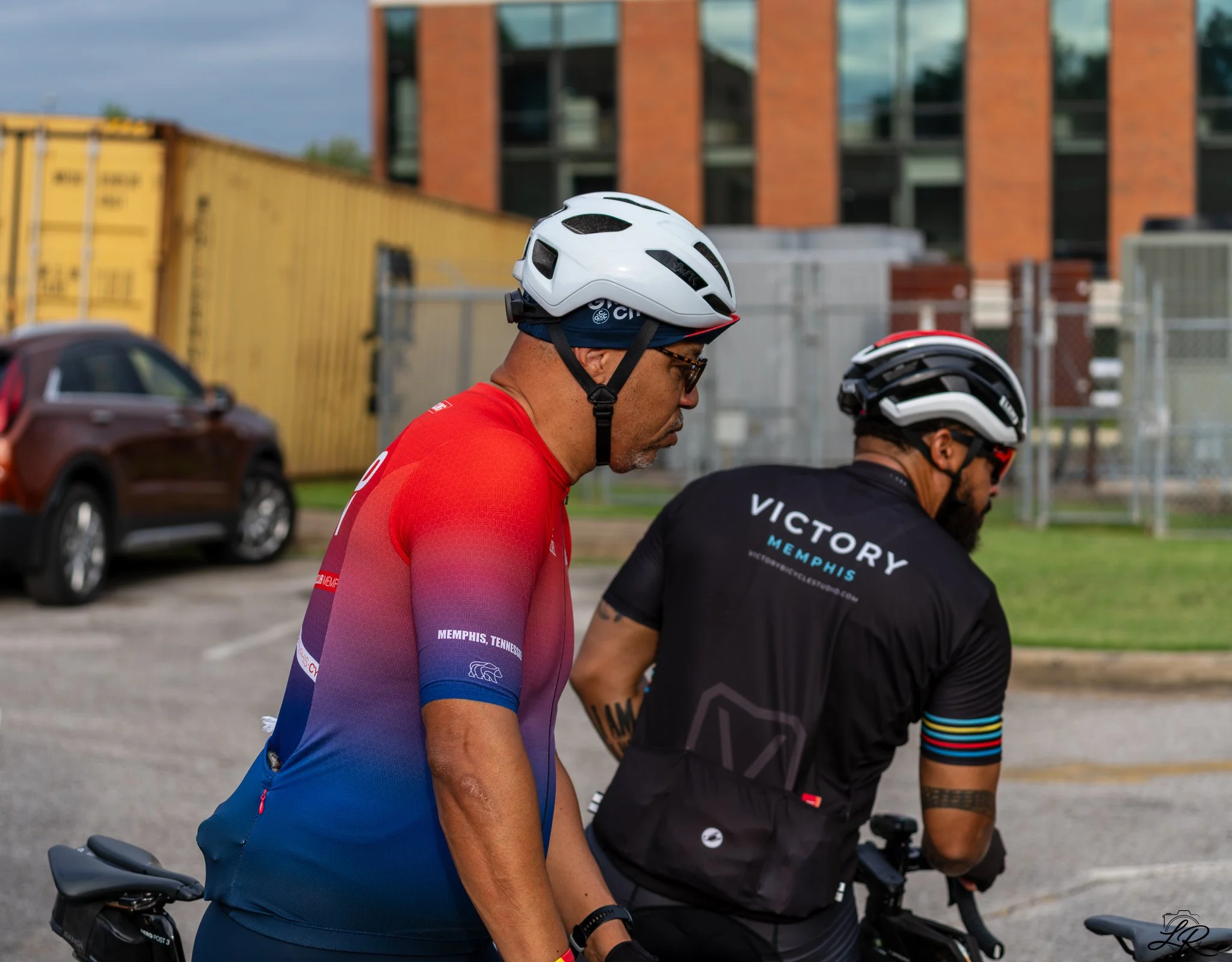 Two cyclists wearing helmets and athletic clothing on their bikes at an outdoor location with cars and buildings in the background.