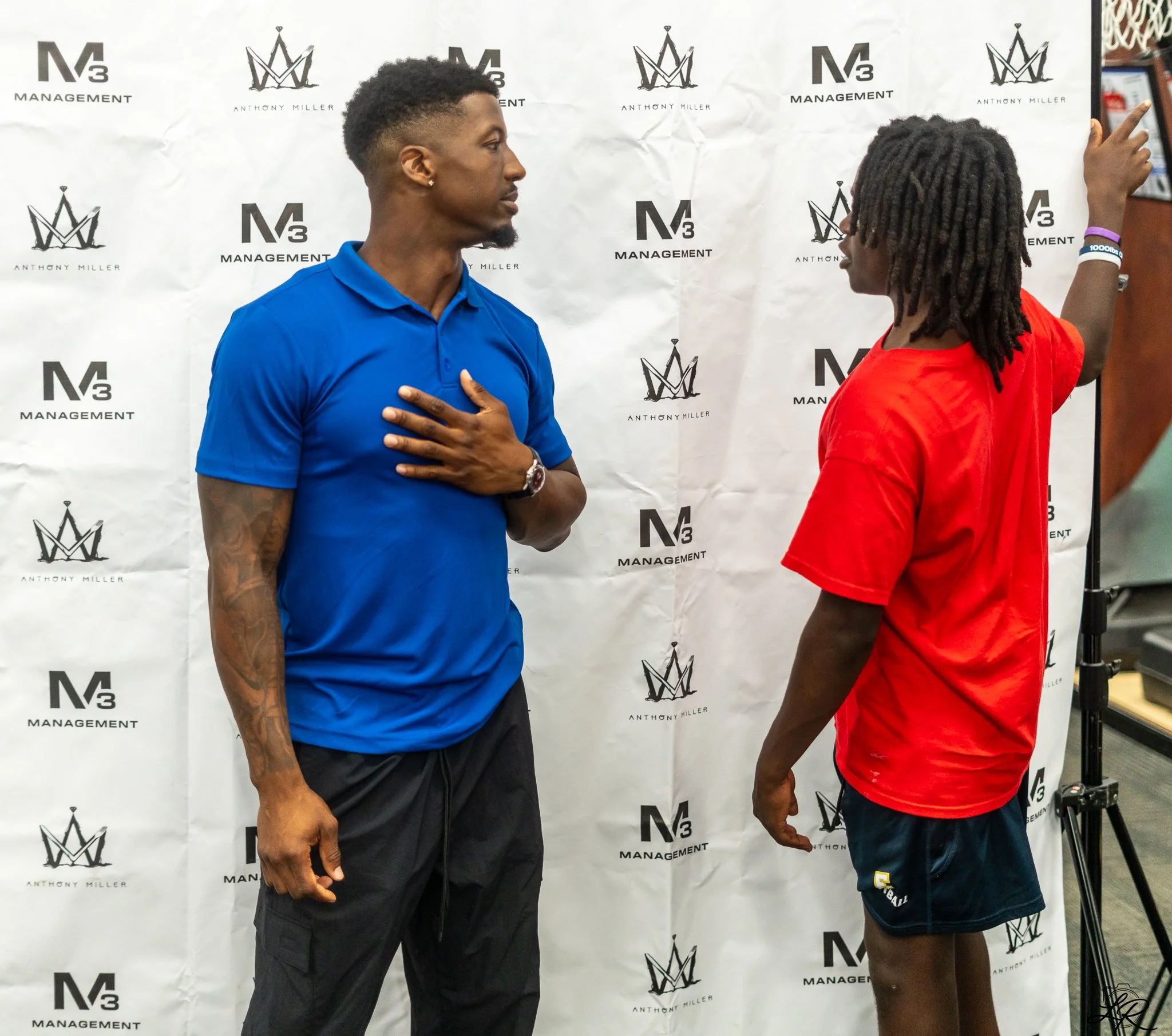 Two men standing in front of a white backdrop with logos, one in a blue polo shirt with his hand on his chest, the other in a red shirt with dreadlocks and wearing a wristband, gesturing with his right hand.
