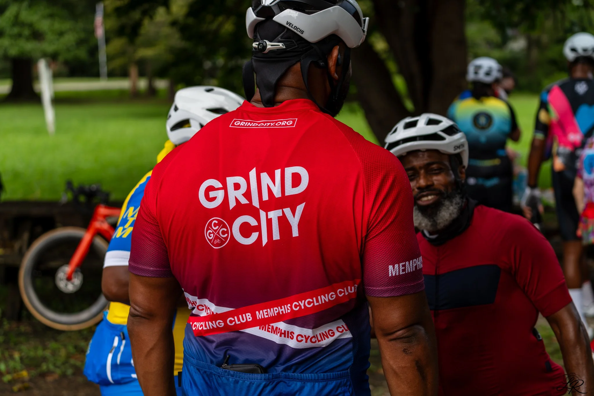 Group of people in cycling gear gathered outdoors, some wearing helmets, with trees and green grass in the background.