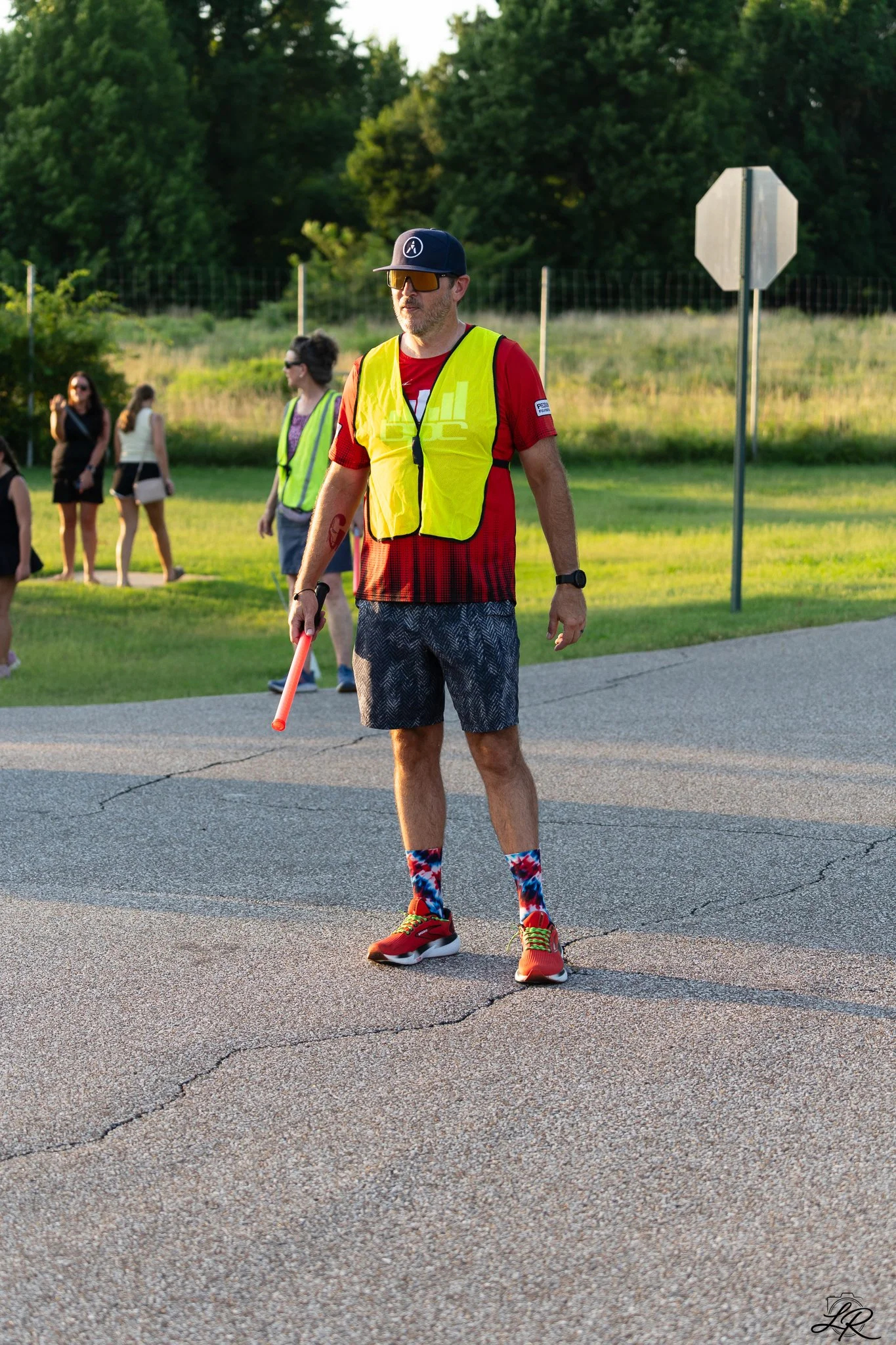 A man wearing a yellow reflective vest, red athletic shirt, black patterned shorts, and colorful socks with red sneakers standing on a paved road at sunset.