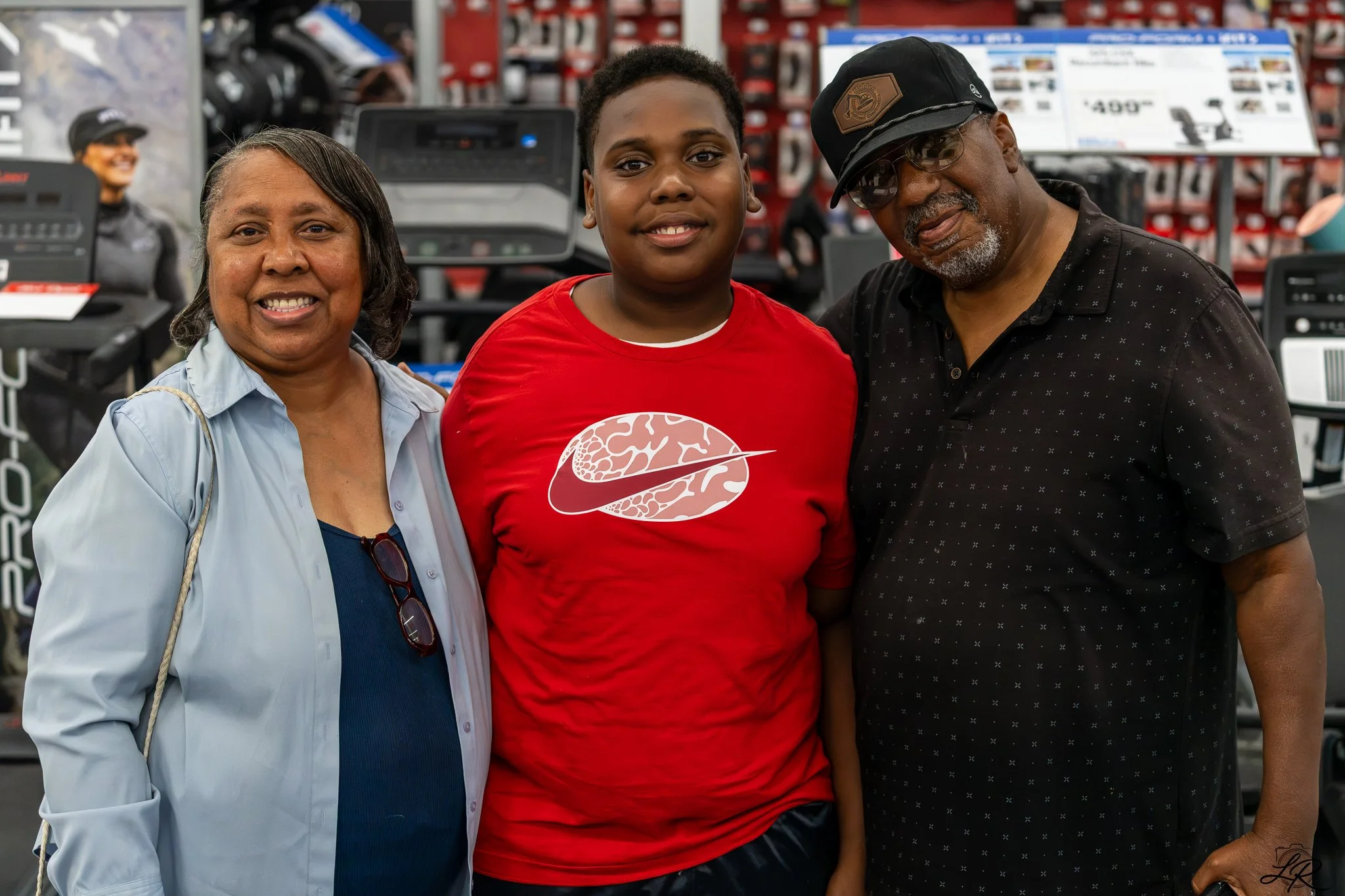 A woman, a young man, and a man standing together inside a store, smiling at the camera. The woman has shoulder-length gray hair, the young man is wearing a red Nike t-shirt, and the man is wearing glasses, a black cap, and a black shirt. There are s