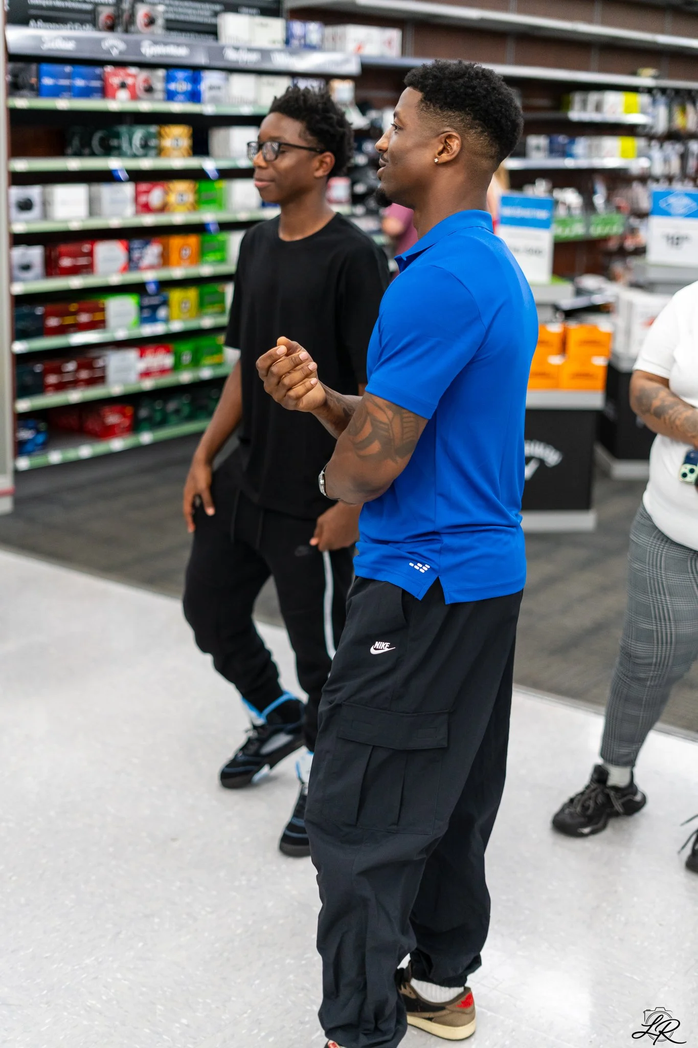 Two young men standing and talking in a store aisle, one wearing a blue shirt and black pants with Nike logo, the other wearing a black shirt and pants, shelves filled with various product boxes in the background.