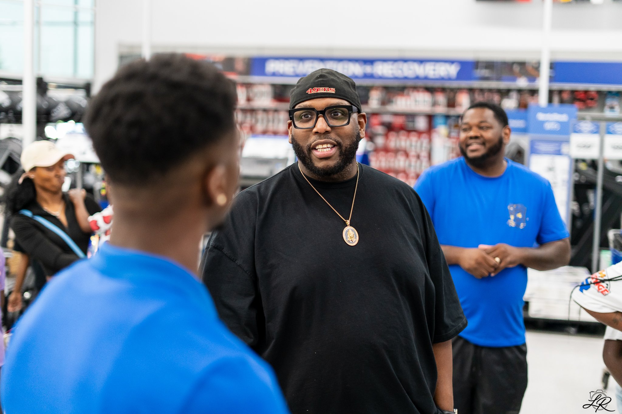 Three men in a store having a conversation, one wearing glasses, a black cap, and a black shirt, while the others wear blue shirts. Shelves with products and a sign indicating 'PREPARATION + RECOVERY' are visible in the background.
