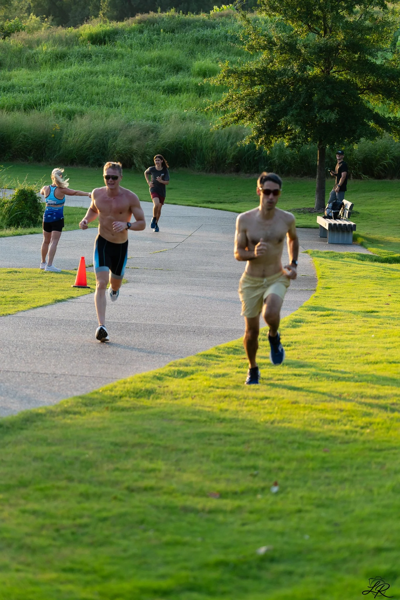 People running on a park pathway with a grassy hill and trees in the background during the late afternoon or early evening.