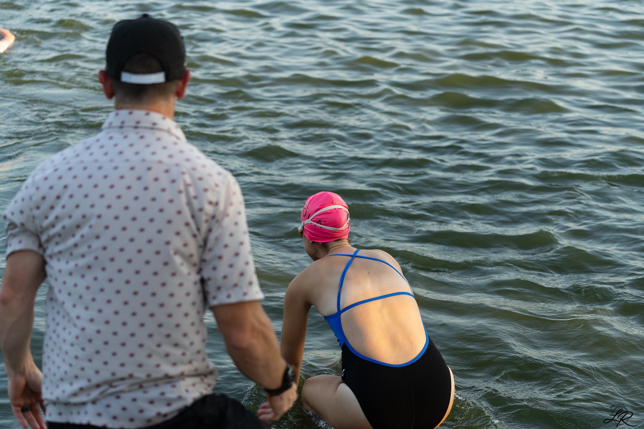 A woman in a pink swim cap and blue swimsuit kneels by the water, while a man in a polka dot shirt and baseball cap stands nearby, with an ocean or lake in the background.