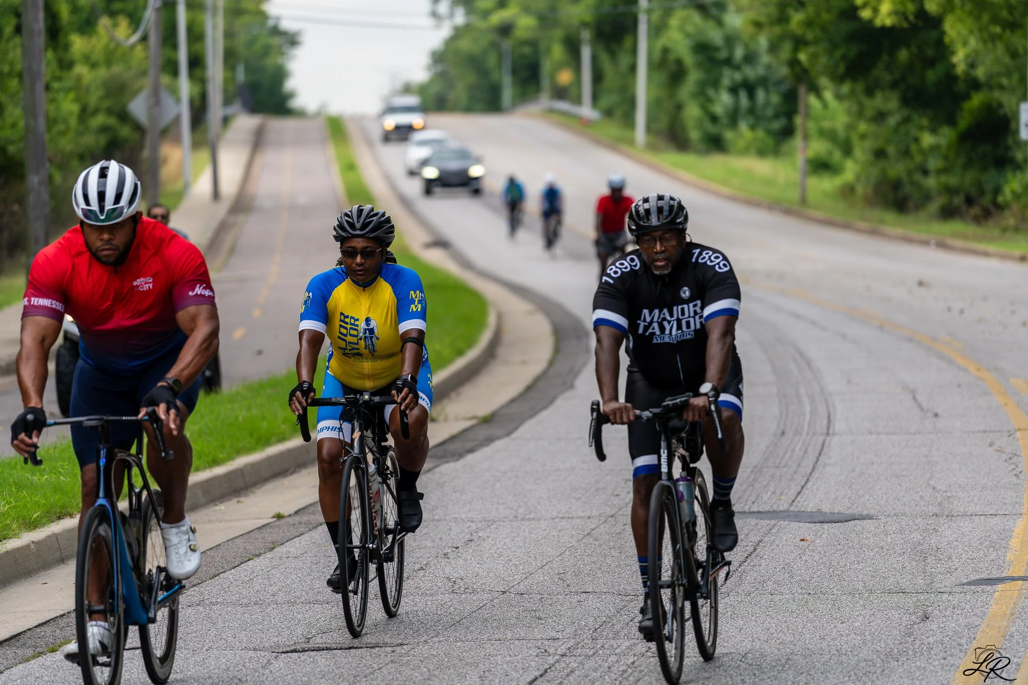 Group of cyclists riding on a suburban road with cars in the background.