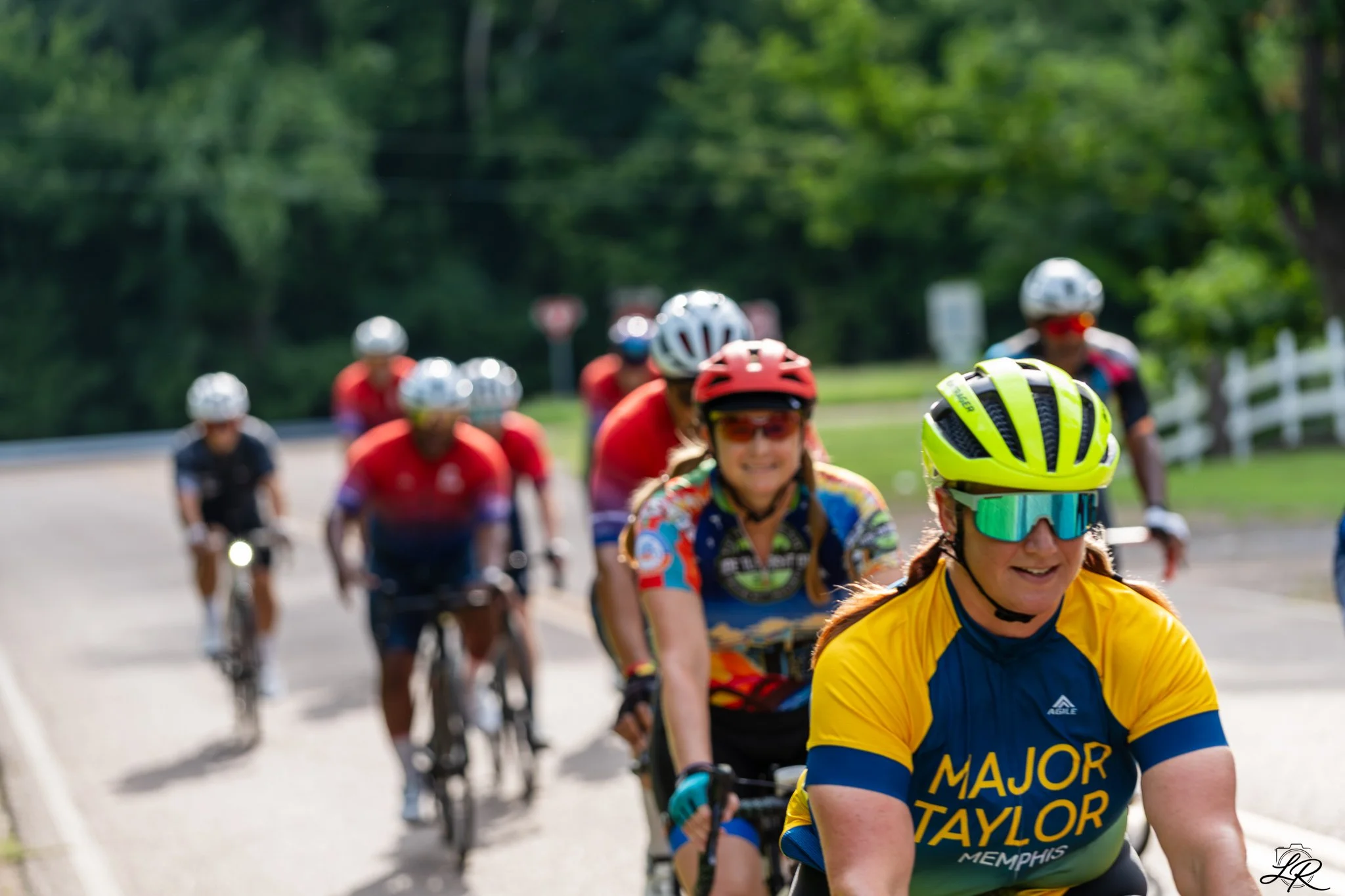 A group of cyclists riding on a road surrounded by green trees, wearing helmets and colorful jerseys, with a focus on a woman smiling in the front wearing a yellow and blue jersey.
