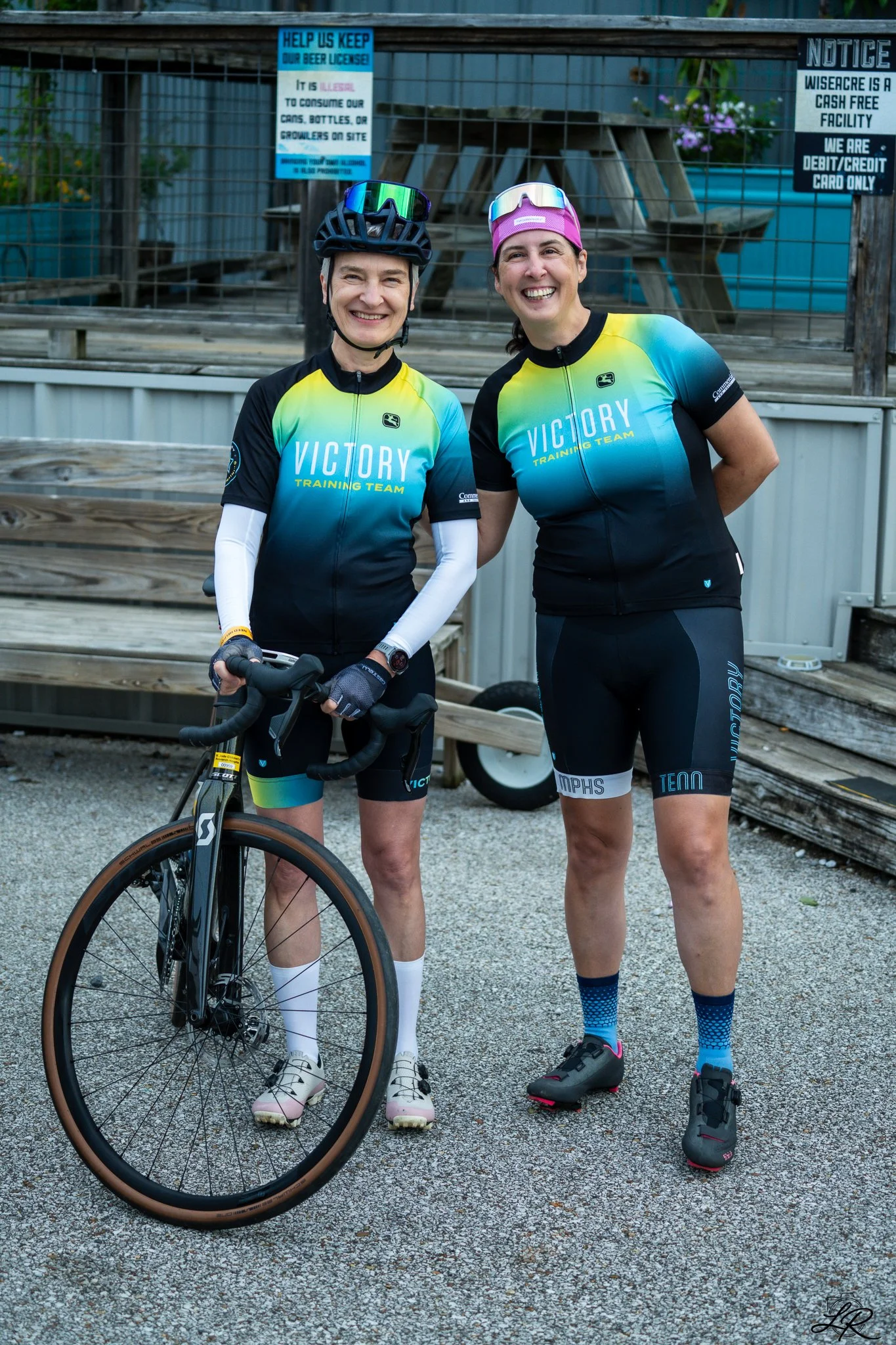 Two women in cycling jerseys with the word "Victory" smiling at the camera. One is holding a bicycle, both wearing helmets, shorts, and cycling shoes.