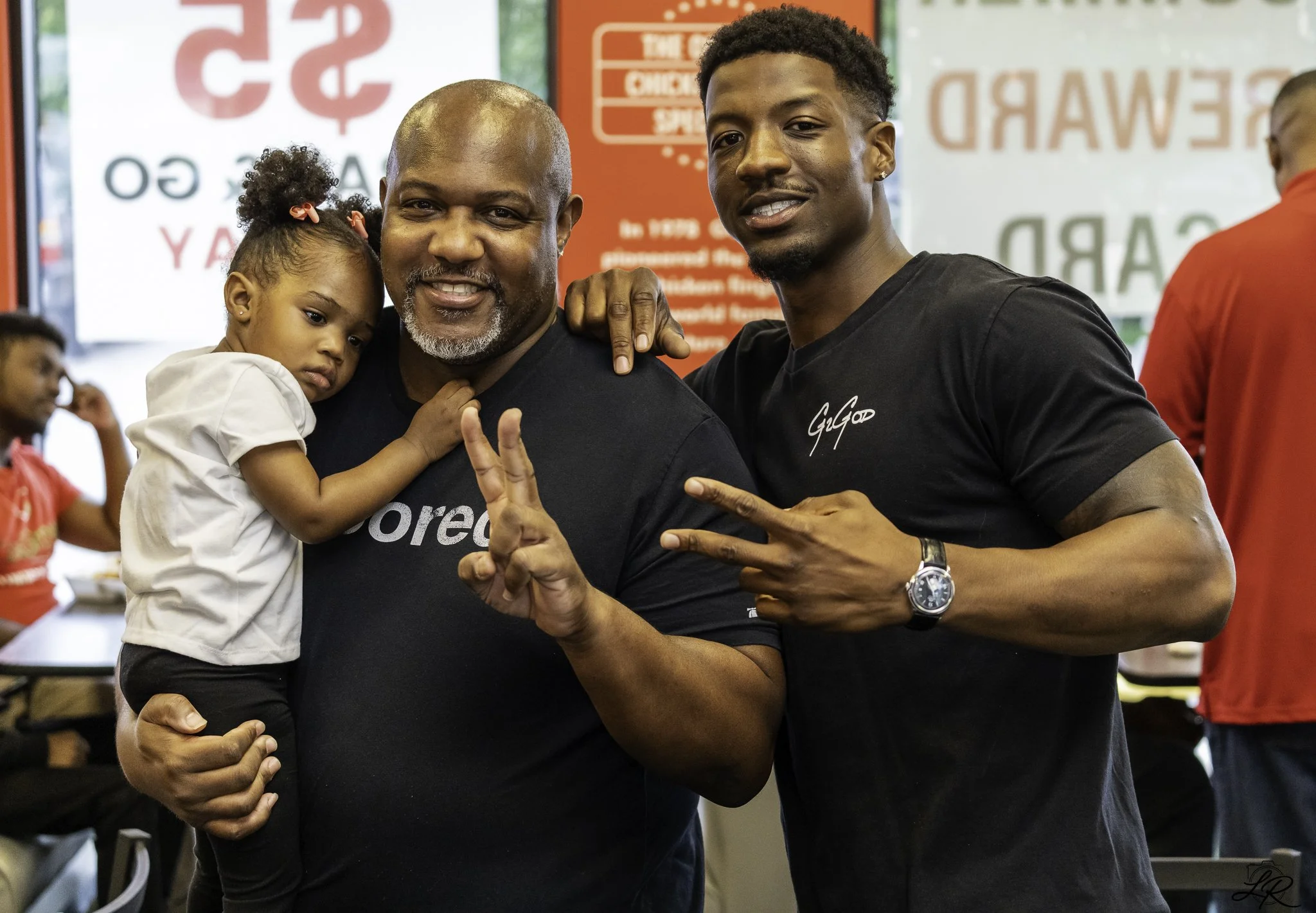 Two men and a young girl pose together indoors, making peace signs with their hands. The girl leans on one man's shoulder, who has a beard and is smiling. The other man is smiling and has his hand on the first man's shoulder. Behind them, there are b