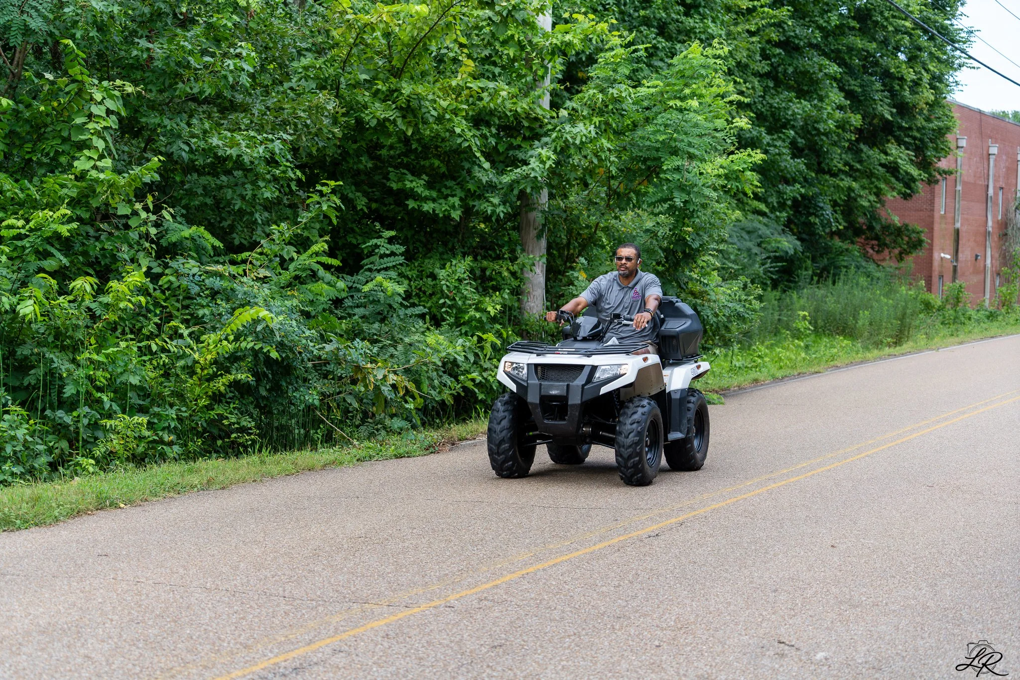 A man riding an all-terrain vehicle (ATV) on a paved road next to a green, leafy area with trees and shrubs, and a red brick building in the background.