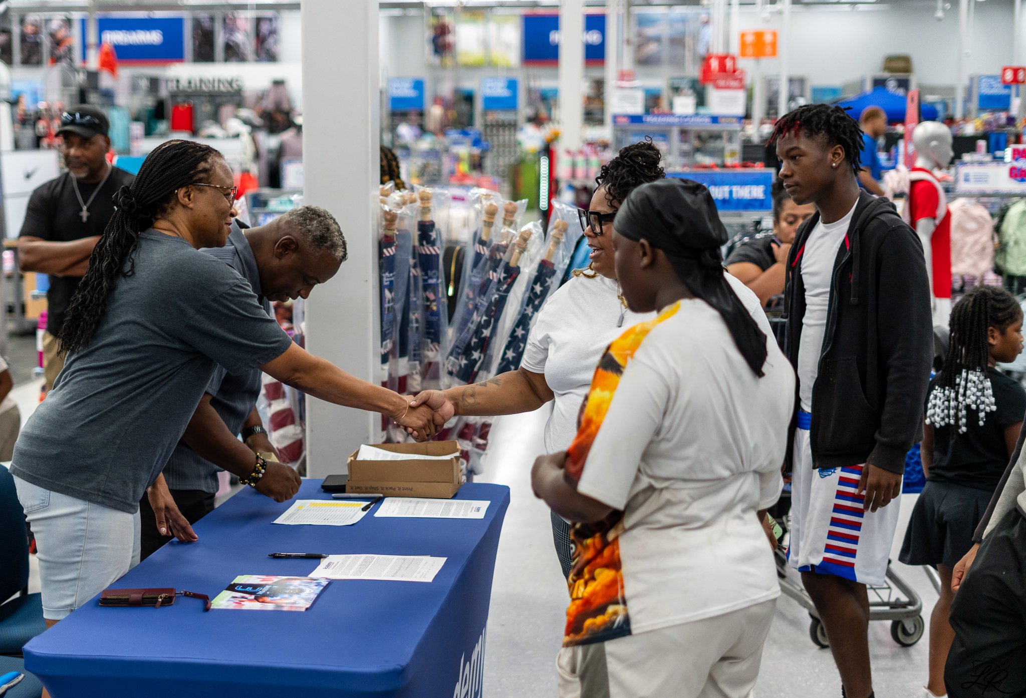 People at a table inside a retail store, engaging in conversation and handshake, with shelves and shopping carts in the background.
