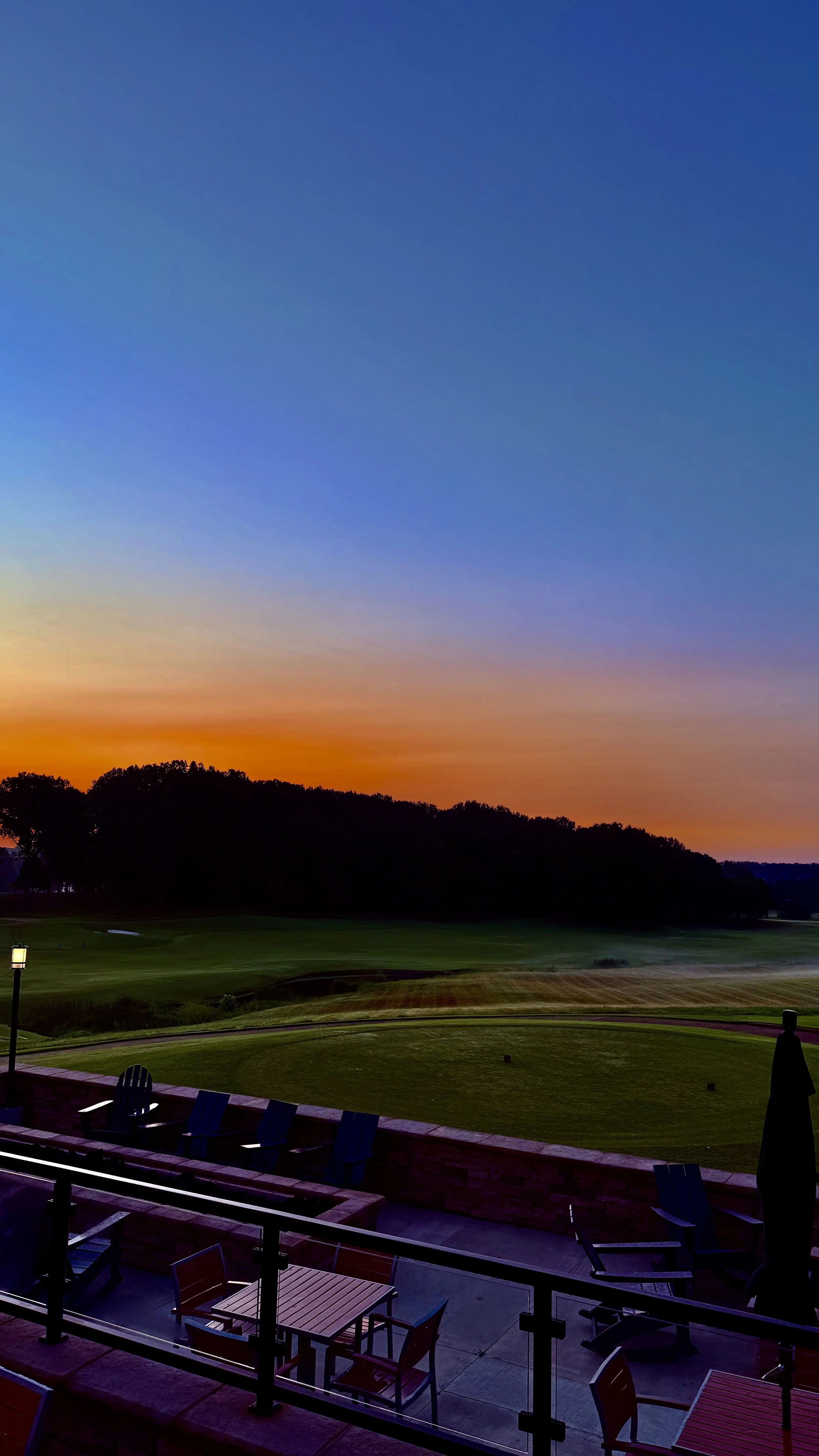 View of a golf course at sunset with a colorful sky, green fairways, and dark trees in the background. There are chairs and tables on a patio with a railing in the foreground.