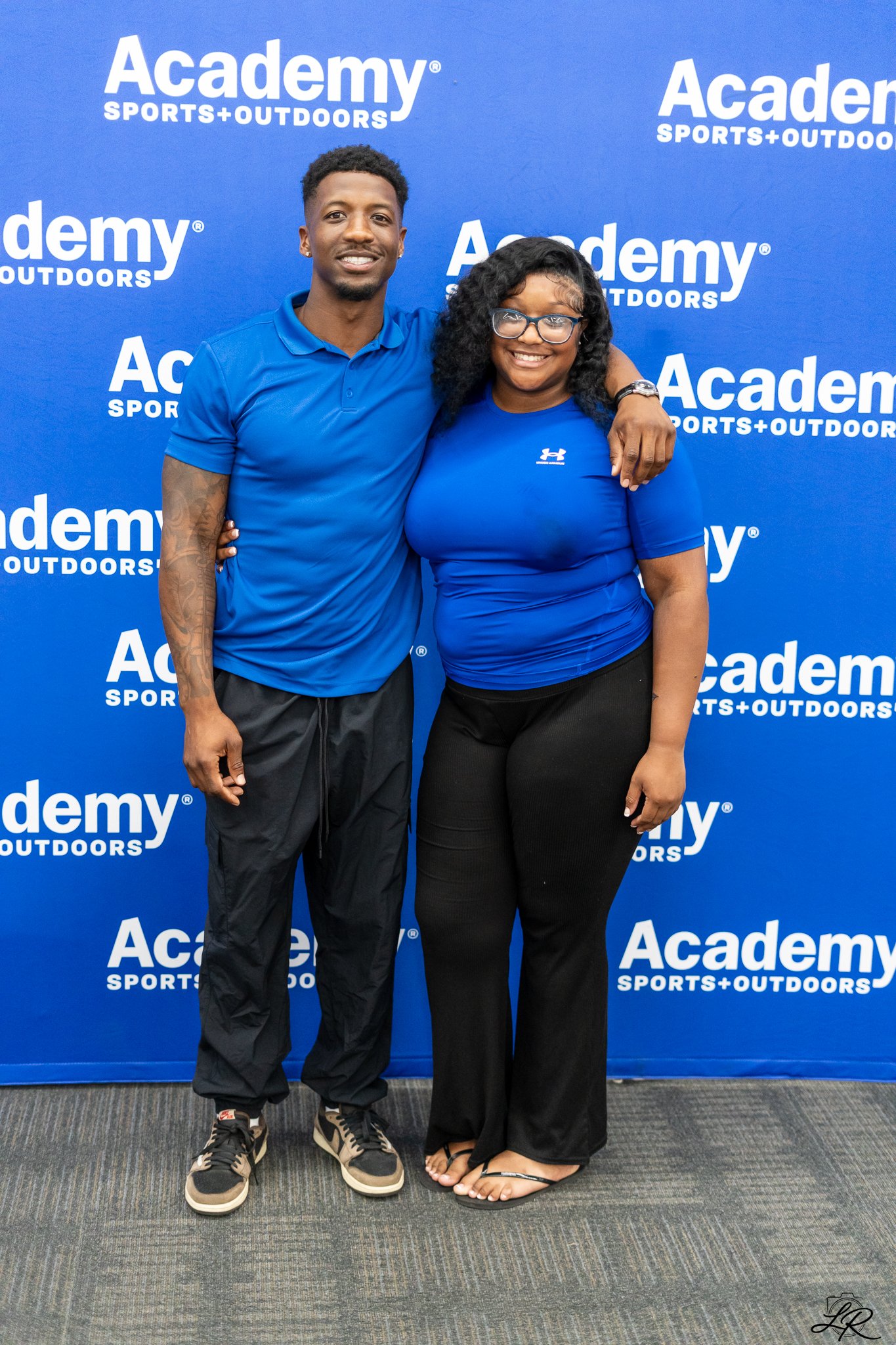 A young man and woman standing together smiling in front of a blue backdrop with the Academy Sports+Outdoors logo, with the man wearing a blue polo shirt and sneakers, and the woman wearing a blue Under Armour shirt, black pants, and flip-flops, with