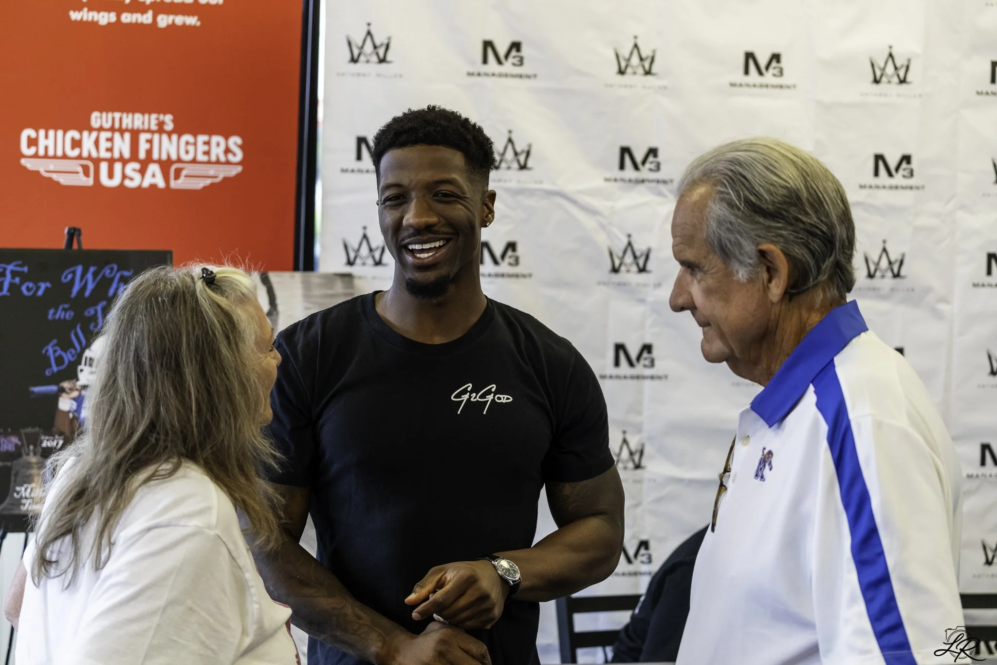 Three people engaging in a conversation at an event with a red and white backdrop. The woman on the left has long blonde hair, the man in the middle has short black hair and a beard, and the man on the right has gray hair. There are banners behind th