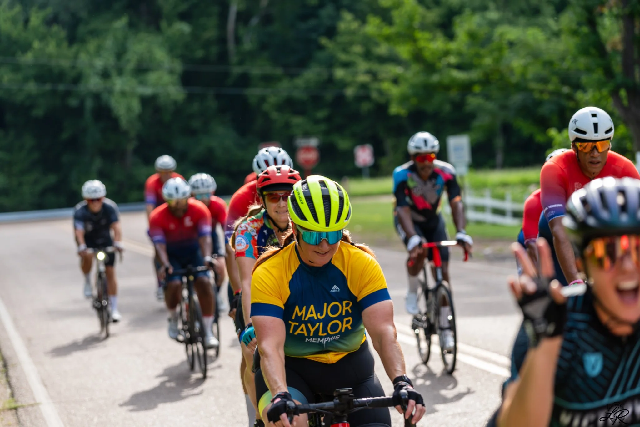 A group of cyclists riding on a rural road with green trees in the background. The woman in the foreground is wearing sunglasses, a yellow and blue cycling jersey, and a yellow helmet. The cyclists are wearing colorful gear and helmets.