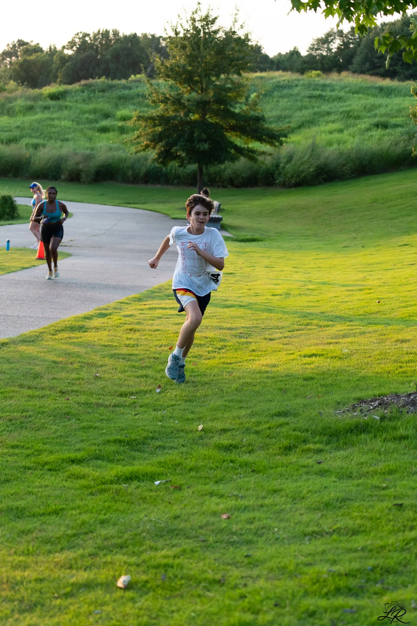A young boy running on a grassy park trail, with a smile on his face, wearing a white t-shirt and shorts. Other people are walking and jogging in the background, surrounded by trees and green hills.