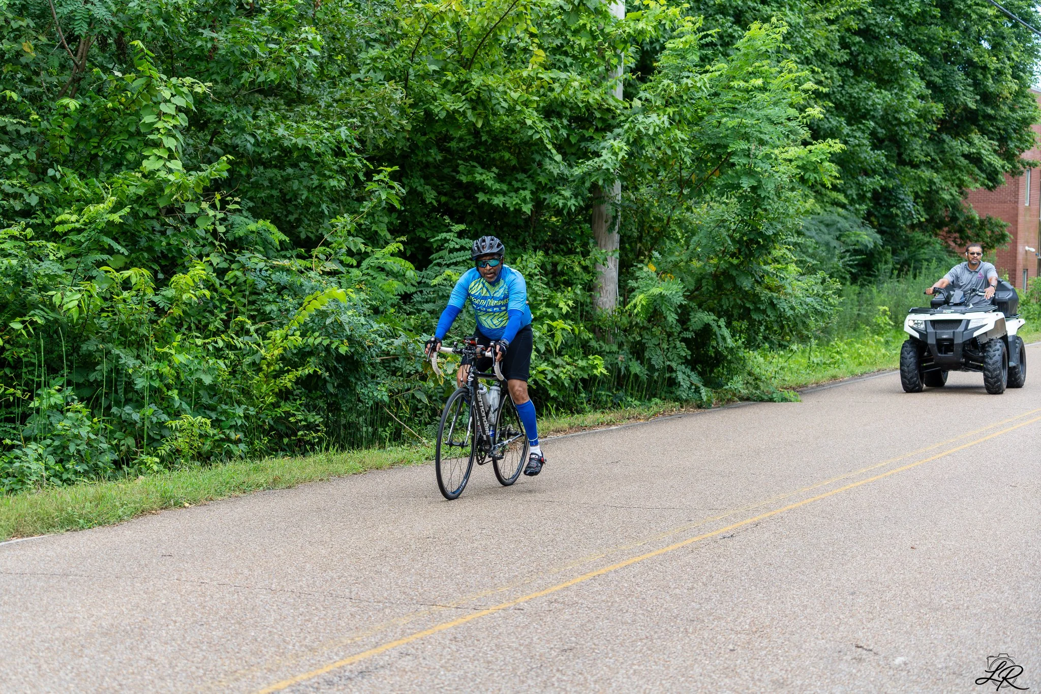 A man riding a bicycle on a paved road beside a large green leafy hedge, with another man on an ATV following behind.