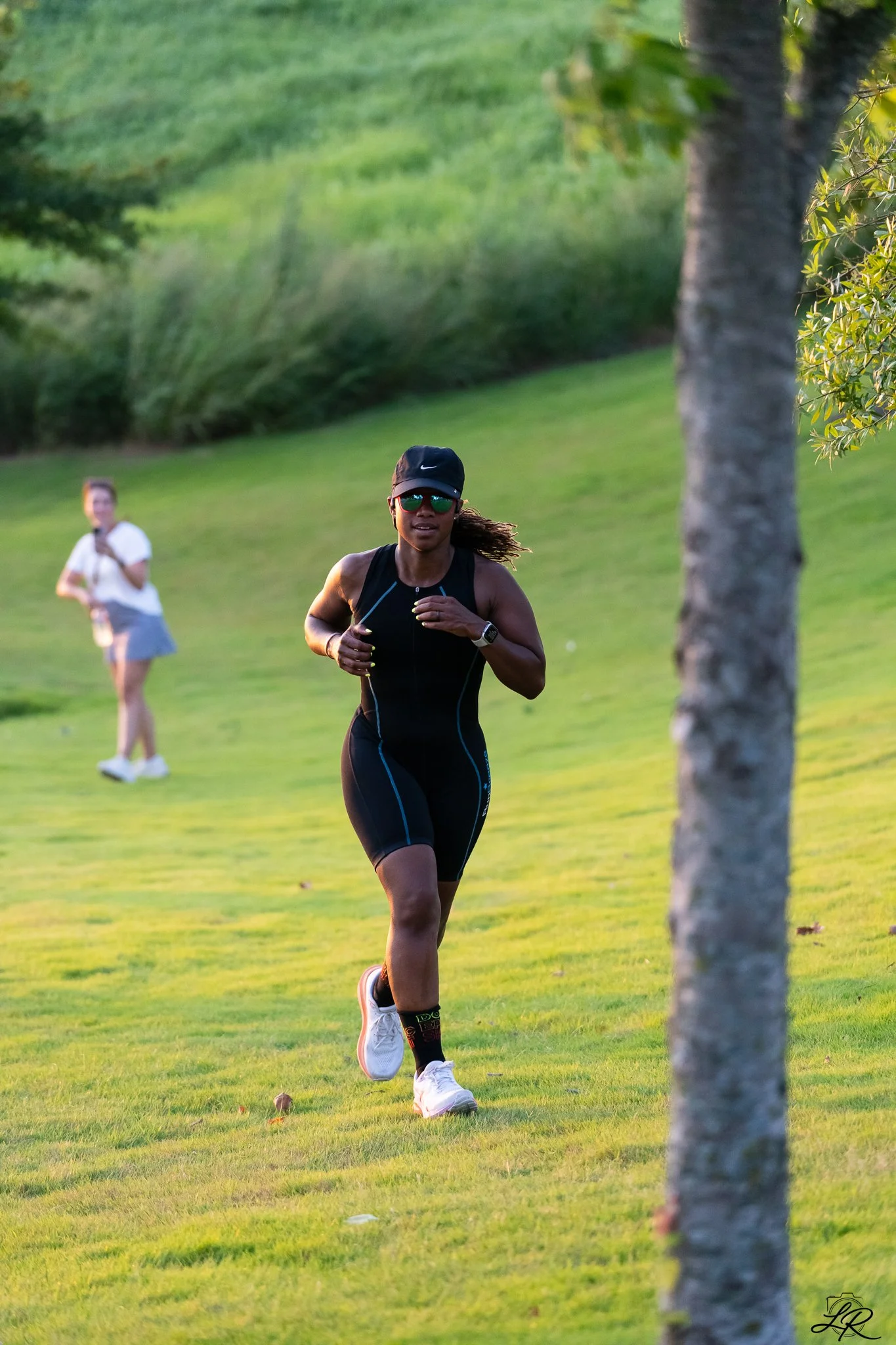 A woman is running outdoors on a grassy field during sunset. She is wearing a black athletic outfit, a black cap, sunglasses, and a fitness tracker, with a pair of white running shoes. There is a tree trunk in the foreground on the right, and another