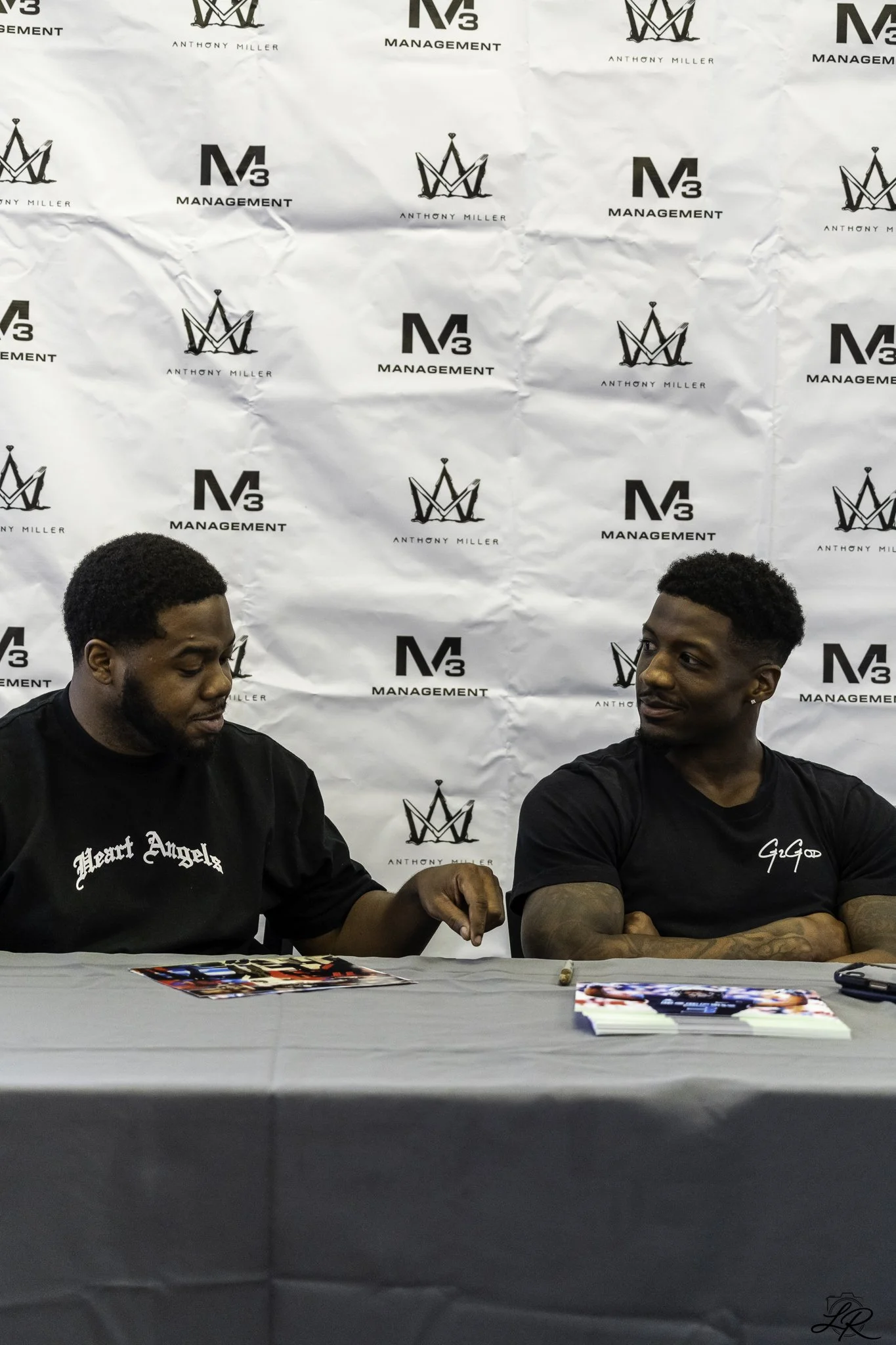 Two men sitting at a table with a gray tablecloth, signing autographs or promotional materials, in front of a backdrop with logos and the name "Anthony Miller". The left man has black hair, a T-shirt with the words "Heart Angels," and a beard. The ri