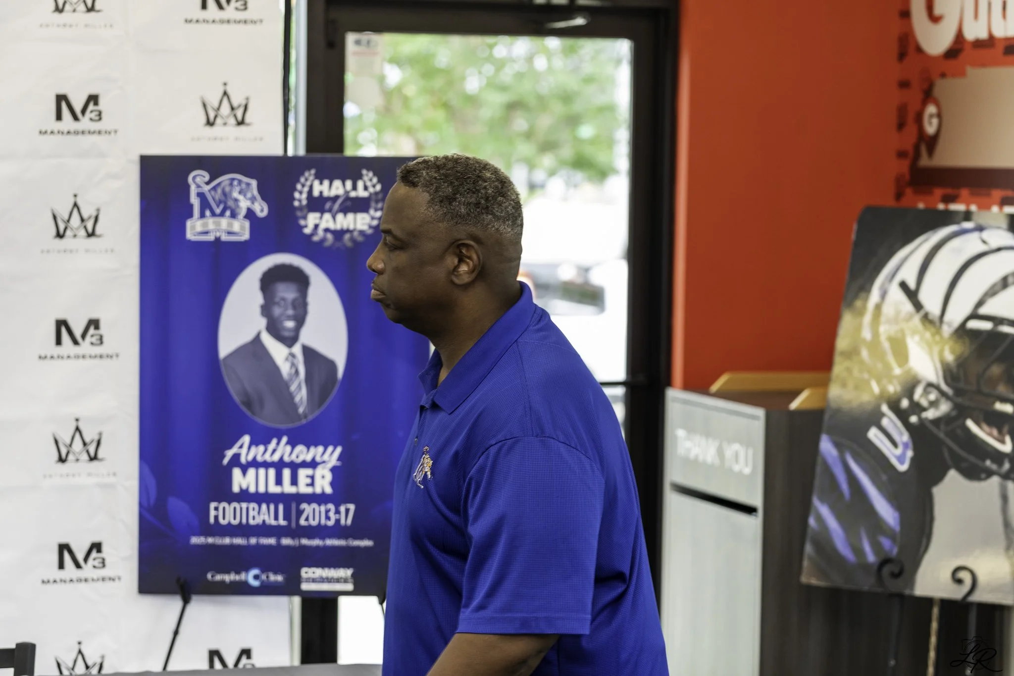 A man in a blue shirt is standing indoors in front of a blue poster with the name Anthony Miller and football information. The background includes a white wall with black logo patterns and a red wall with some posters and artwork.