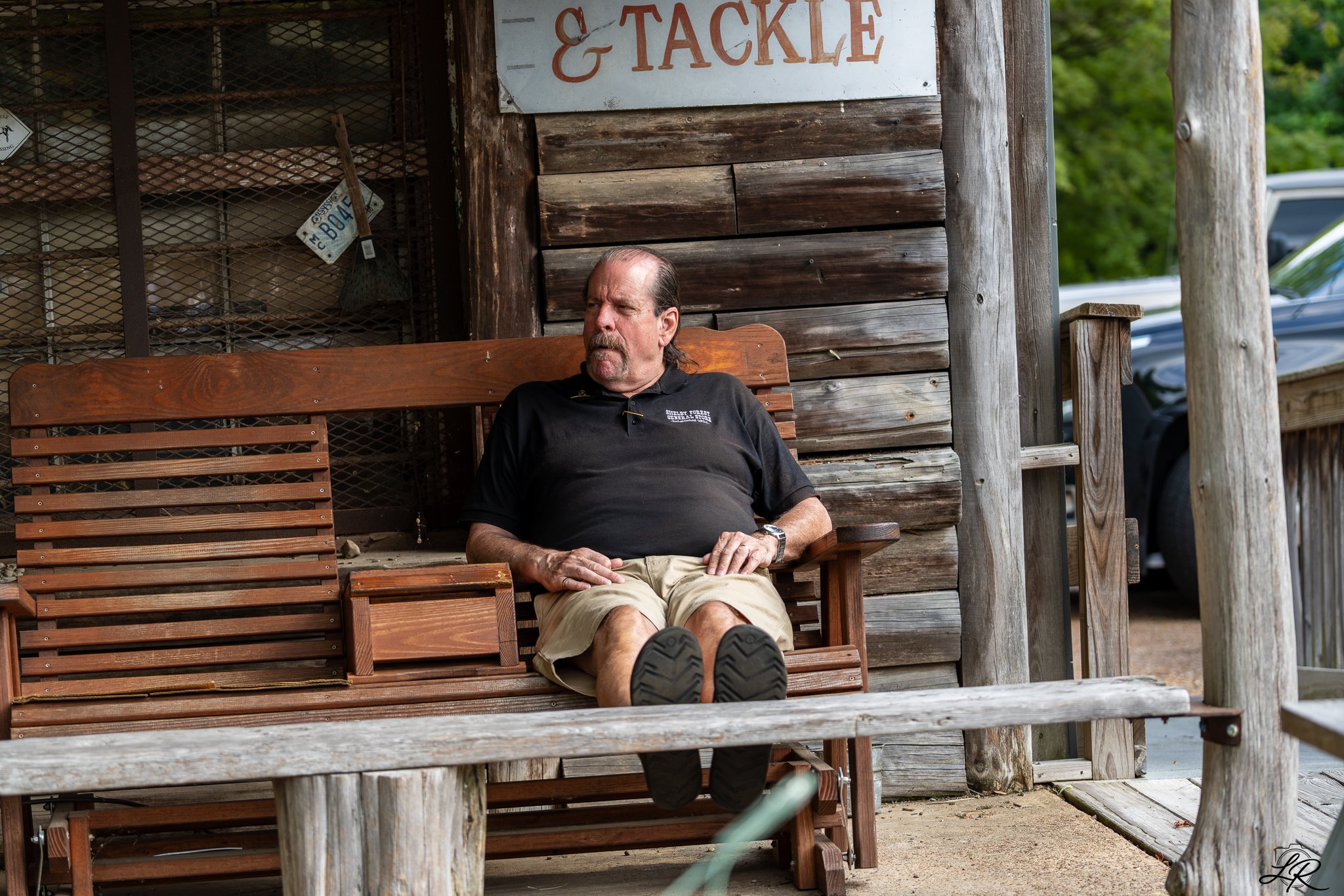 A man sitting on a wooden bench outside a rustic building with a sign that says 'Tackle'. He is wearing a black shirt and khaki shorts, with his legs extended and shoes visible in the foreground. Green trees and parked cars are in the background.