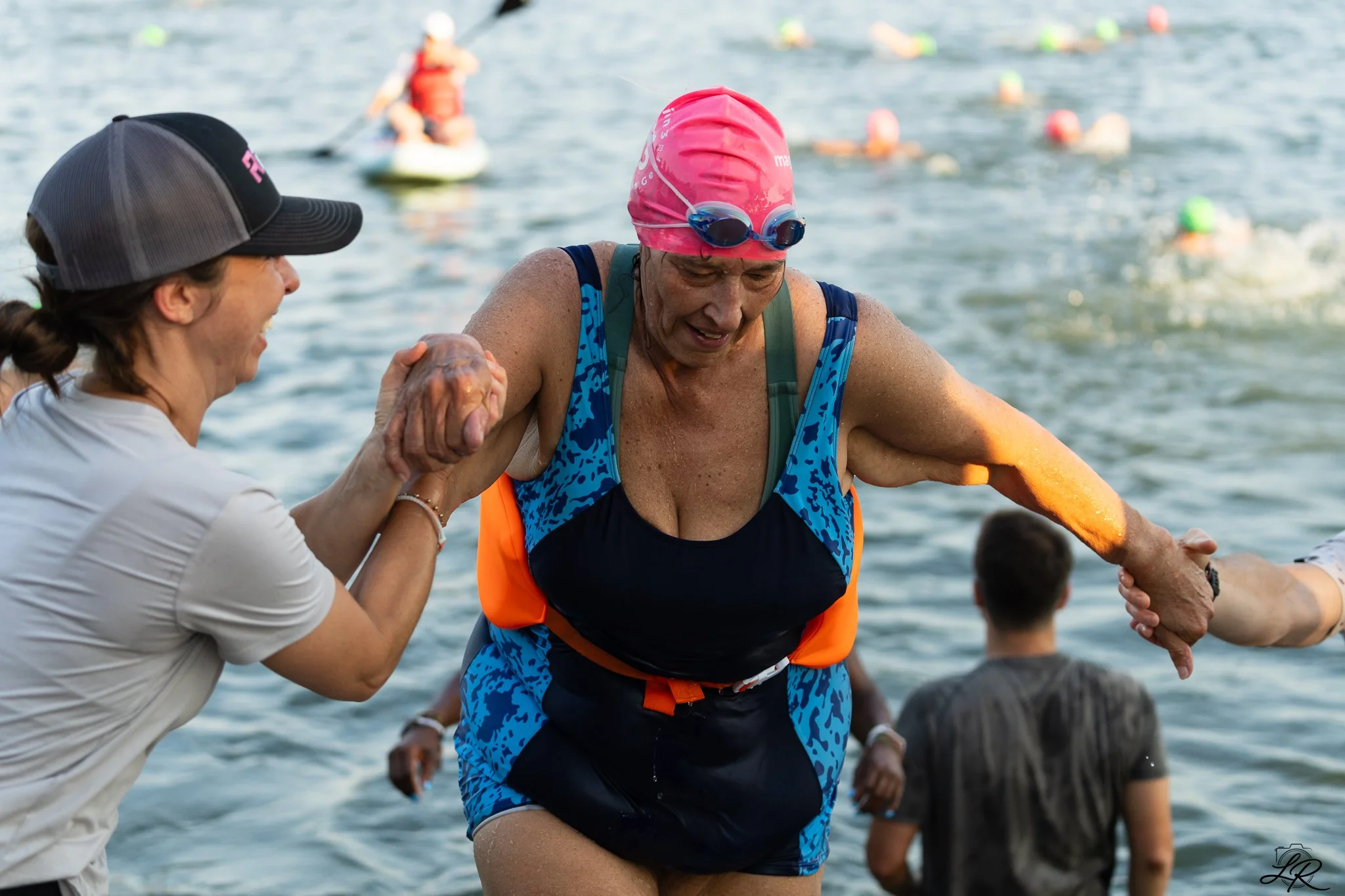 A woman with a pink swim cap and goggles is being helped out of the water by another woman in a white shirt and black baseball cap during a triathlon or open water swimming event, with other swimmers in the background.