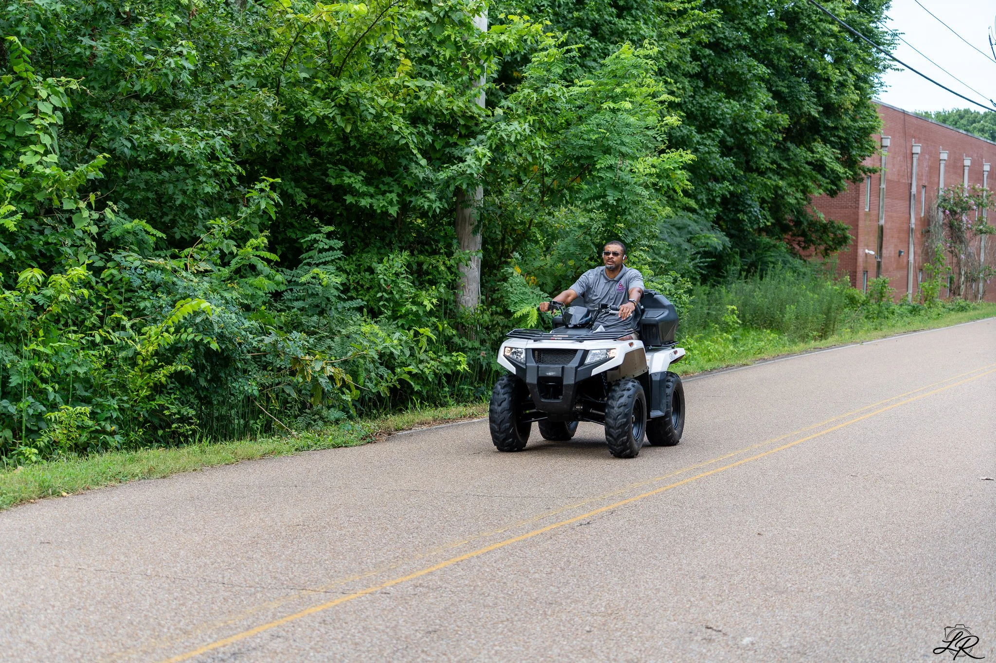 A person riding an all-terrain vehicle (ATV) on a paved road near green trees and a brick building.
