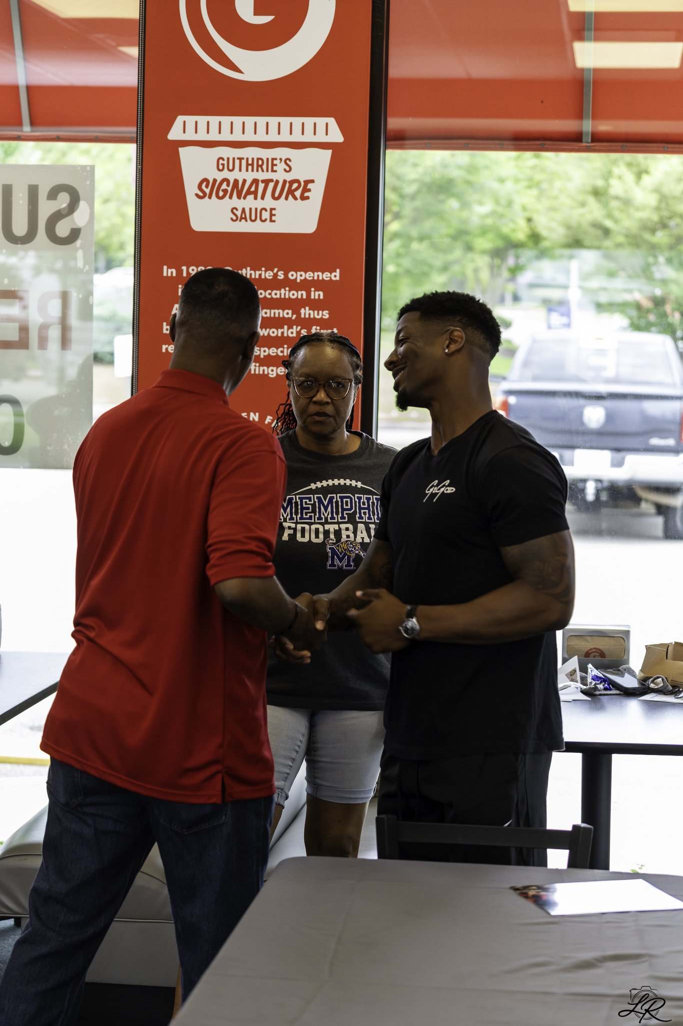 Three people standing indoors, two men shaking hands and a woman observing. A red sign in the background advertises 'Guthrie's Signature Sauce' with additional text about the sauce's history. The setting appears to be a casual dining or event area wi