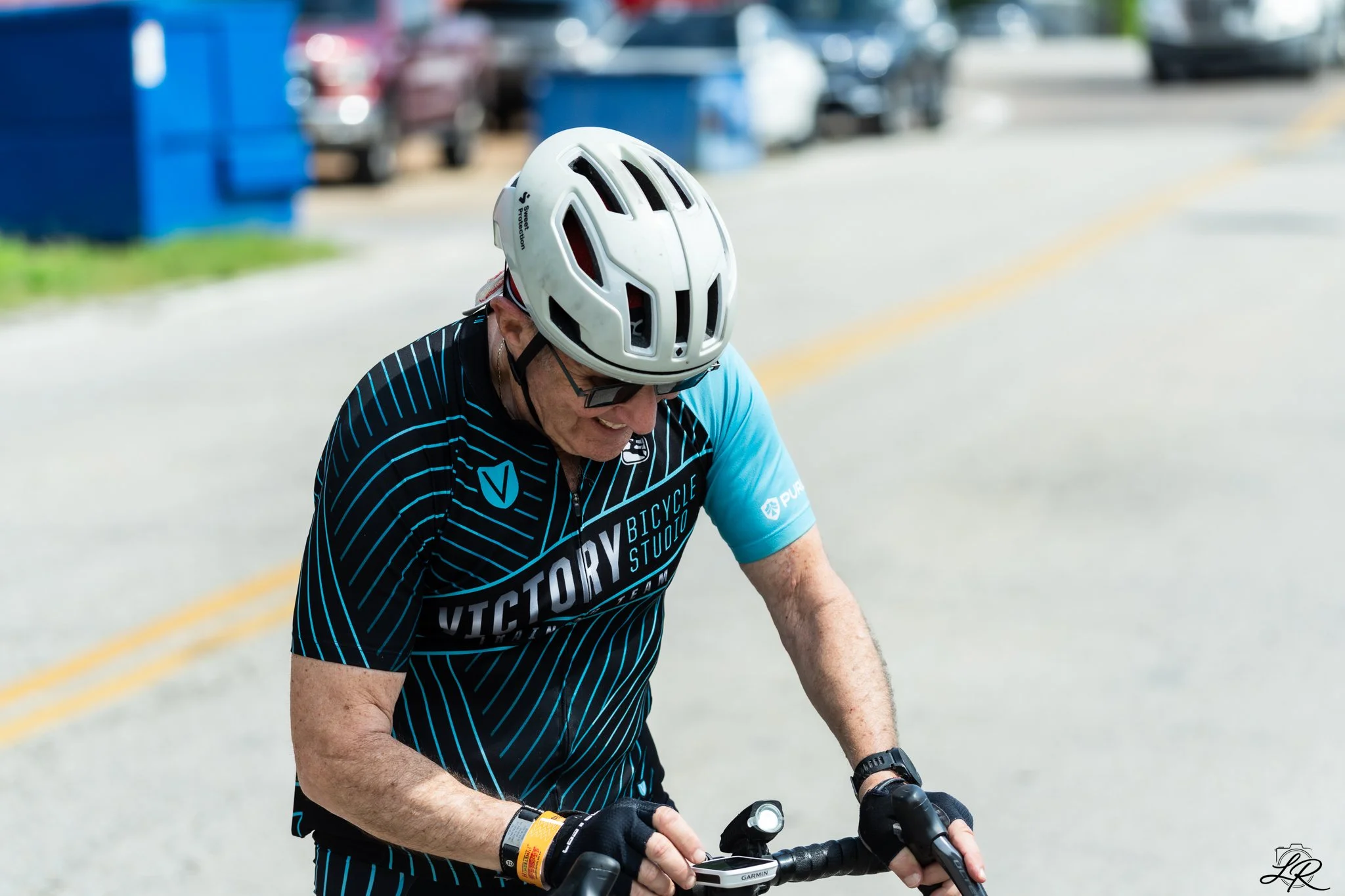 A person wearing a bicycle helmet and sunglasses riding a bicycle on a paved road. The person is wearing a black and blue cycling jersey with the word 'Victory' on it, and appears to be smiling or enjoying themselves.