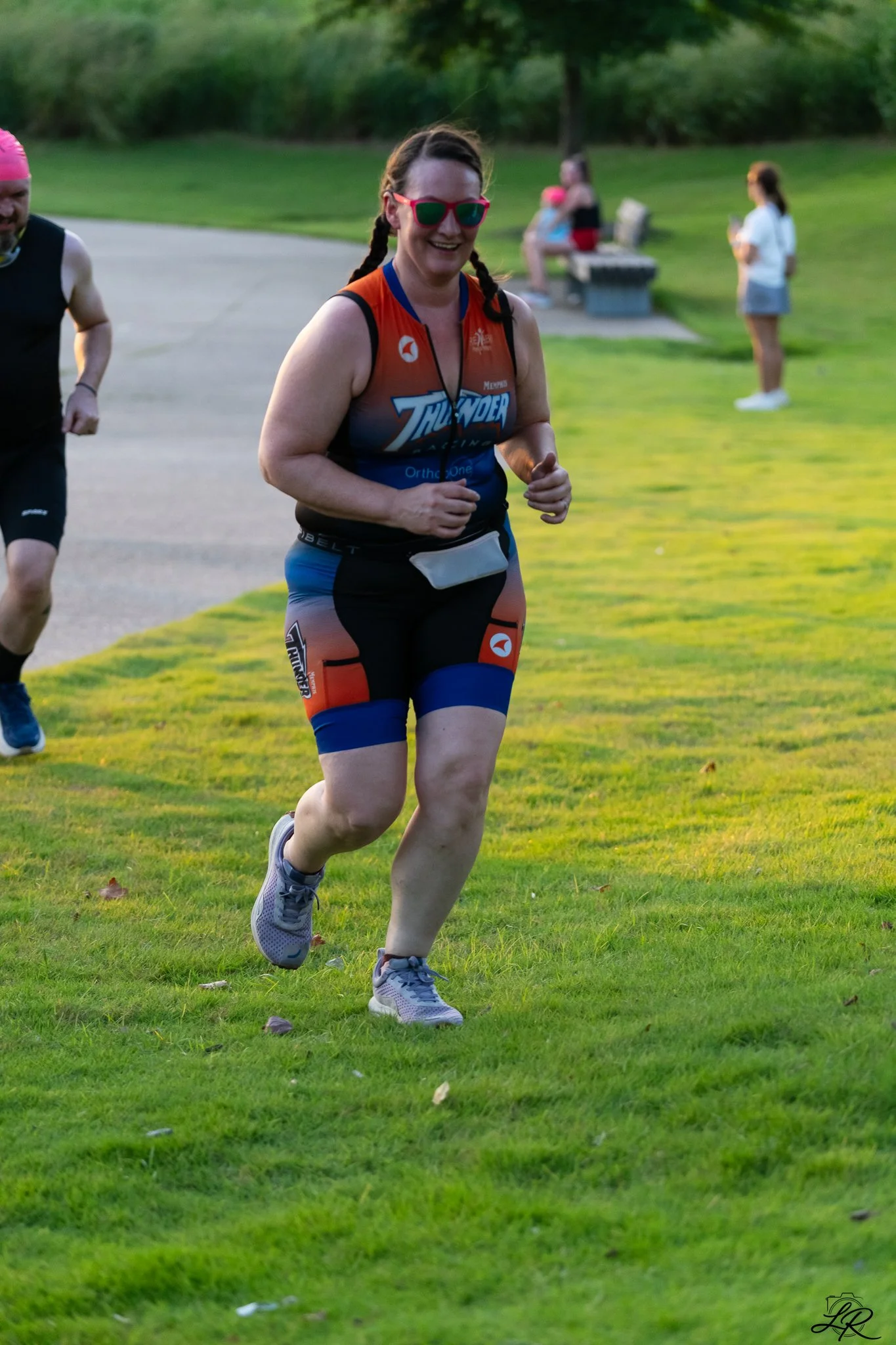 A woman running outdoors on grass, wearing colorful athletic gear and sunglasses, smiling. In the background, other people are sitting on benches.
