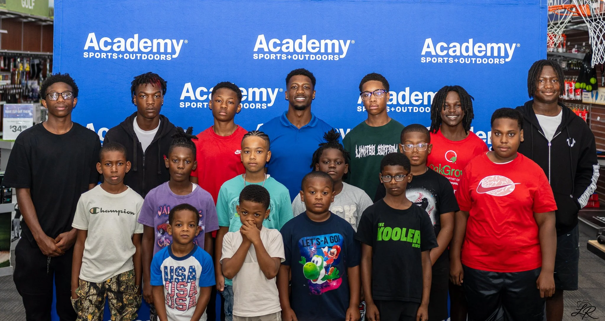 A group of children and teenagers standing together in front of a blue backdrop with 'Academy Sports + Outdoors' logos, inside a retail store.