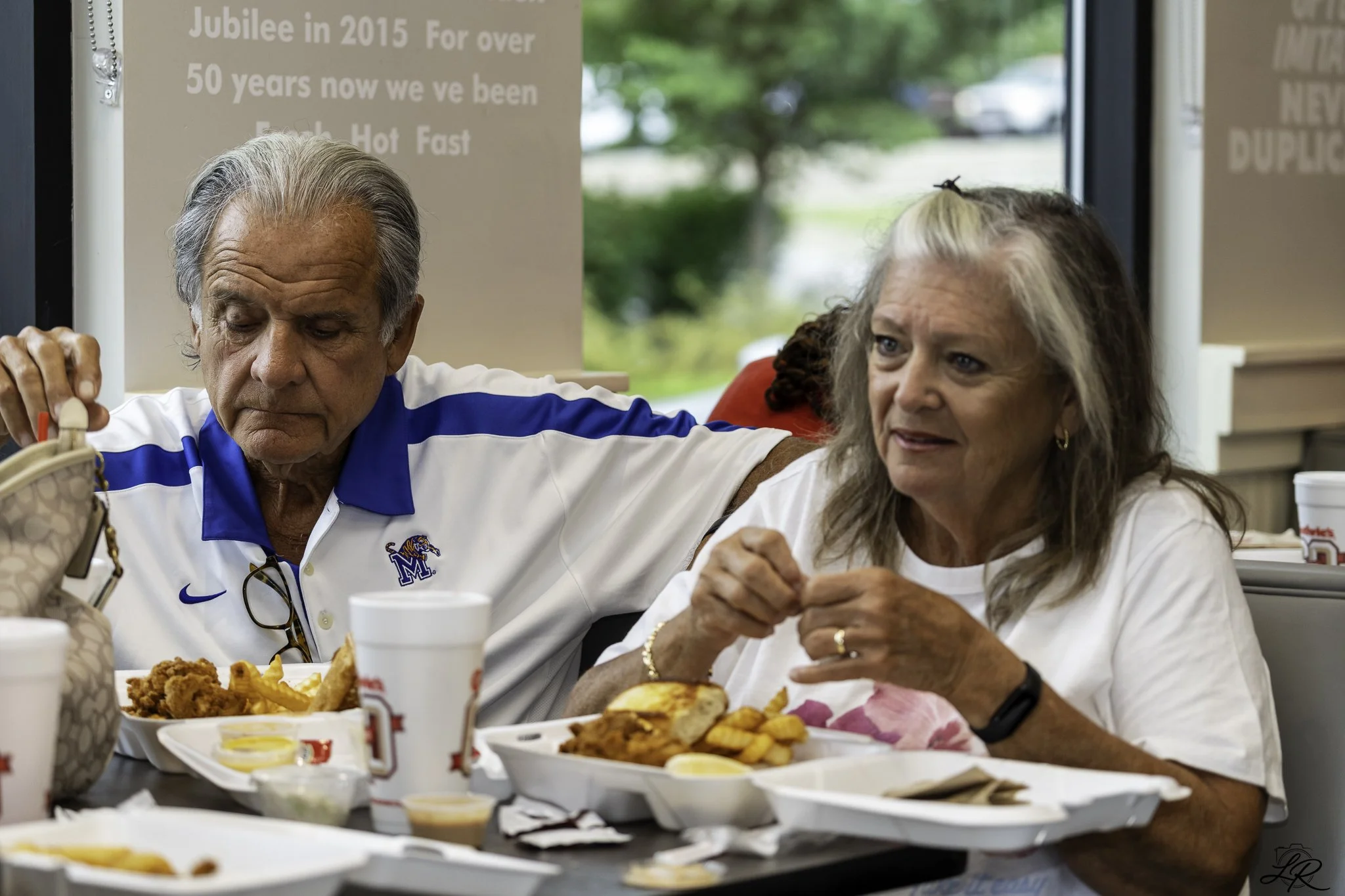 An elderly man in a white and blue sports polo and an elderly woman in a white top are sitting at a table, enjoying a meal together inside a restaurant. The table has multiple trays of fried foods, French fries, and dipping sauces. The background has