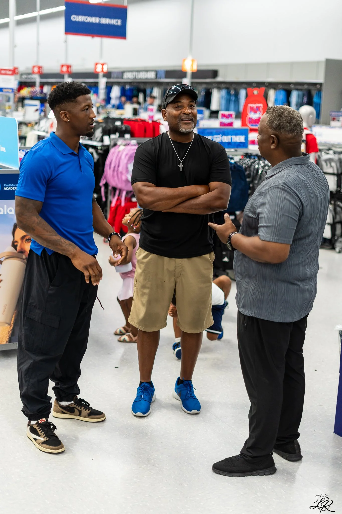 Three men in conversation inside a retail store, with shelves stocked with clothing and signs indicating customer service and checkout in the background.