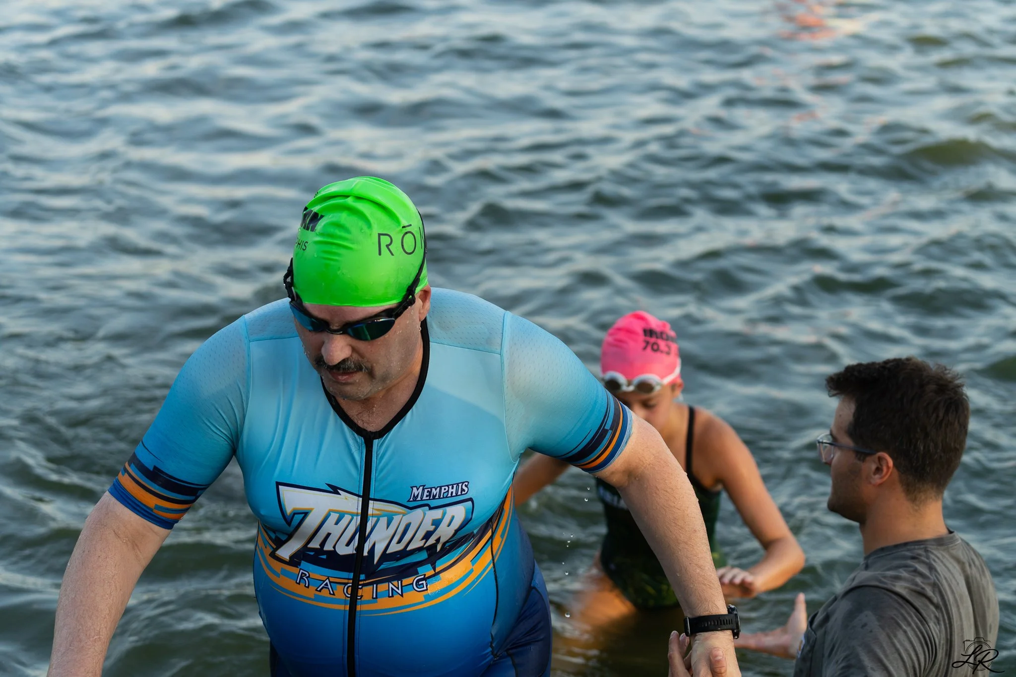 A man wearing a bright green swim cap, sunglasses, and a blue athletic shirt is in a body of water, participating in a race or swim event. Behind him, others are also swimming or exiting the water, with one person wearing a pink swim cap.