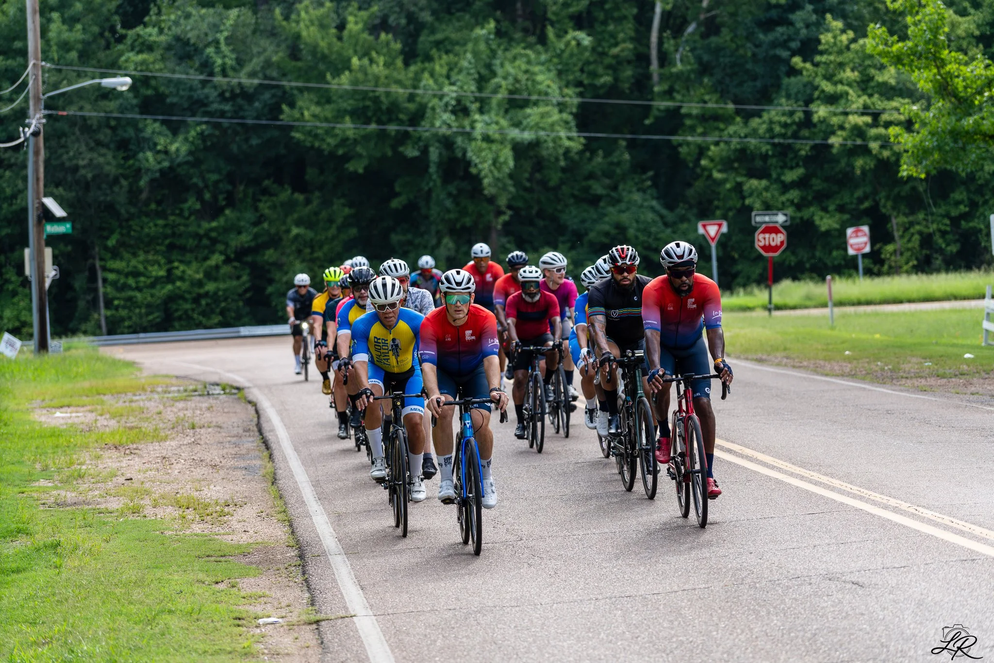 A group of cyclists riding on a rural road surrounded by green trees, with several street signs visible in the background.
