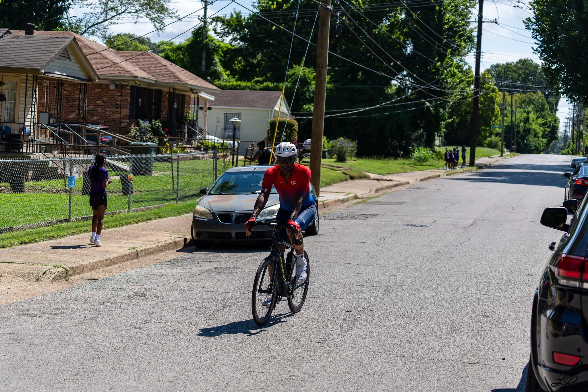 A man wearing a red cycling jersey and a helmet rides a bicycle on a residential street. There are children playing near a house, and a girl stands on the sidewalk watching. The street is lined with parked cars and utility poles with wires.