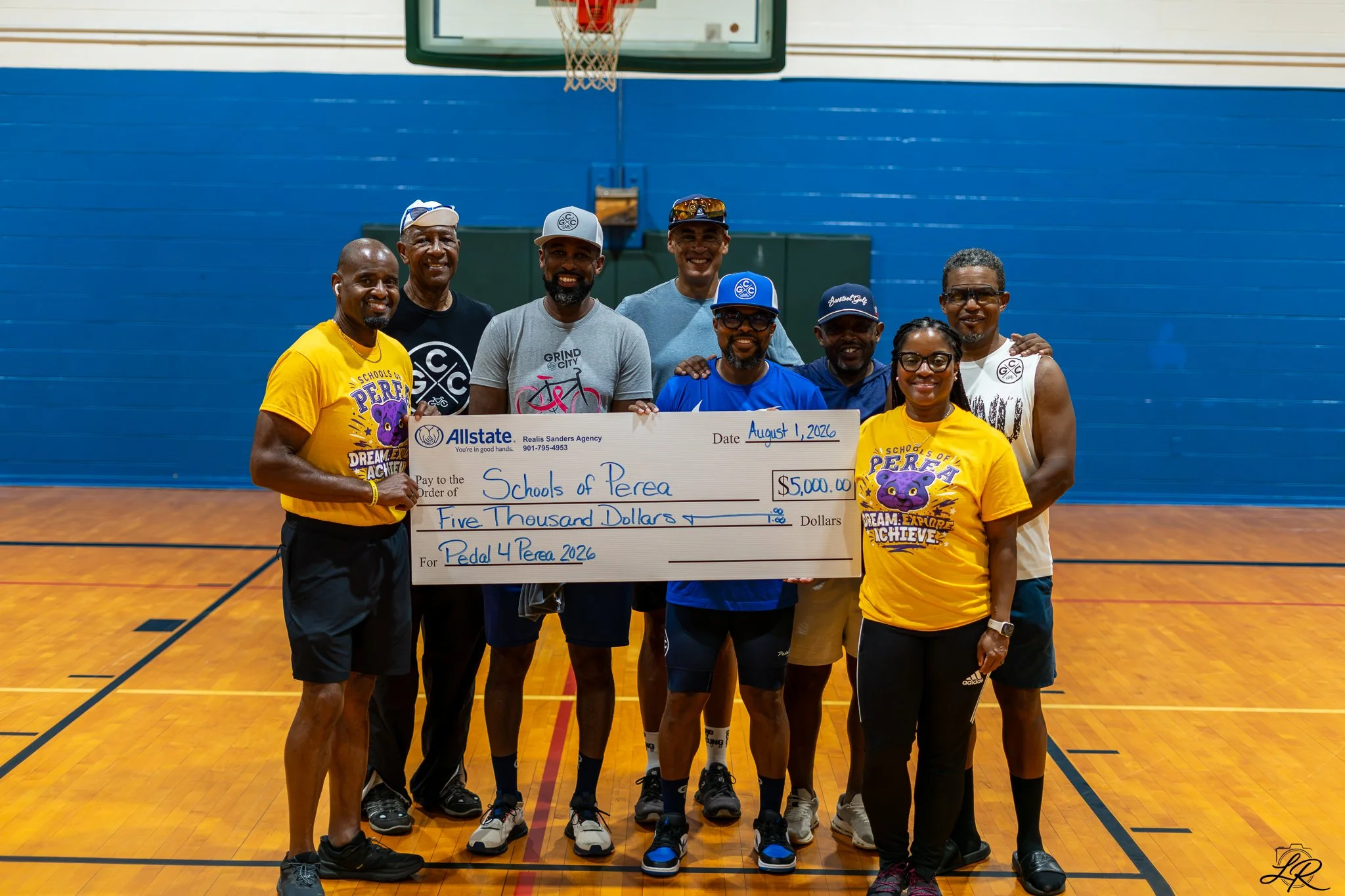Group of eight adults standing on a basketball court holding a large check for $5,000 made out to Schools of Perea, with a basketball hoop and blue wall in the background.