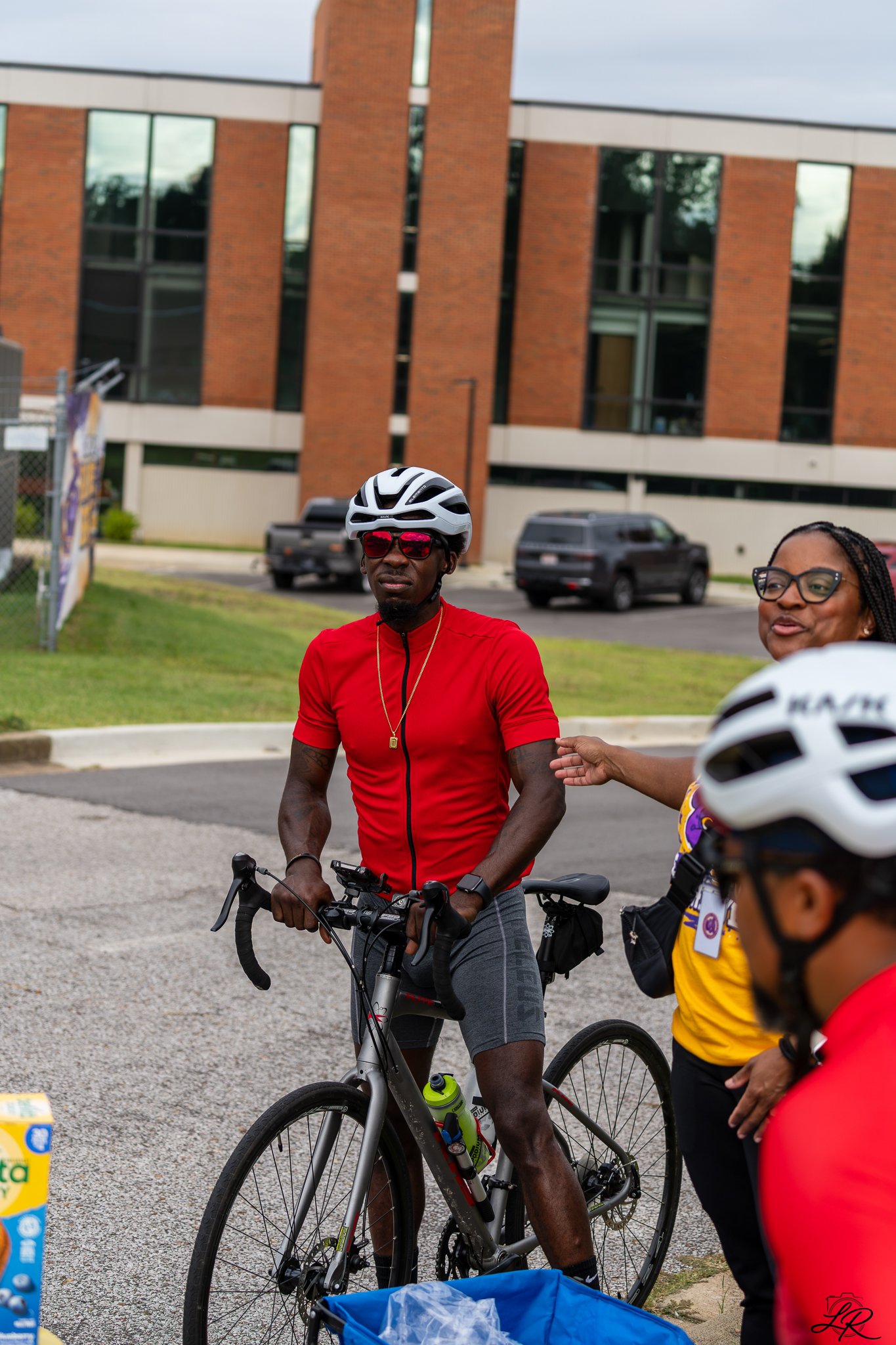 Three people with bicycles standing on a sidewalk near a brick building, wearing helmets and casual athletic clothing, engaged in conversation.