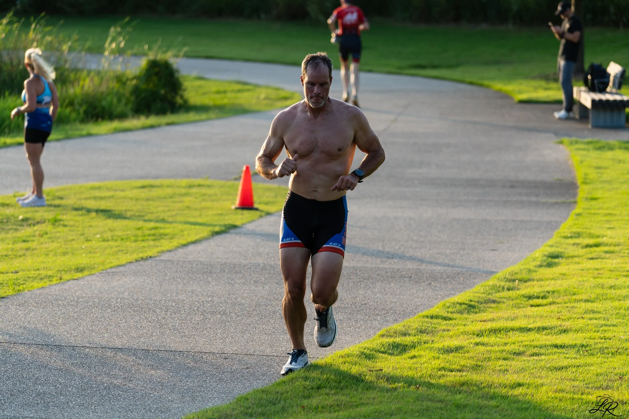 A man running shirtless outdoors on a paved pathway with green grass on the side, during daylight. In the background, three people are standing or sitting, some looking at their phones.