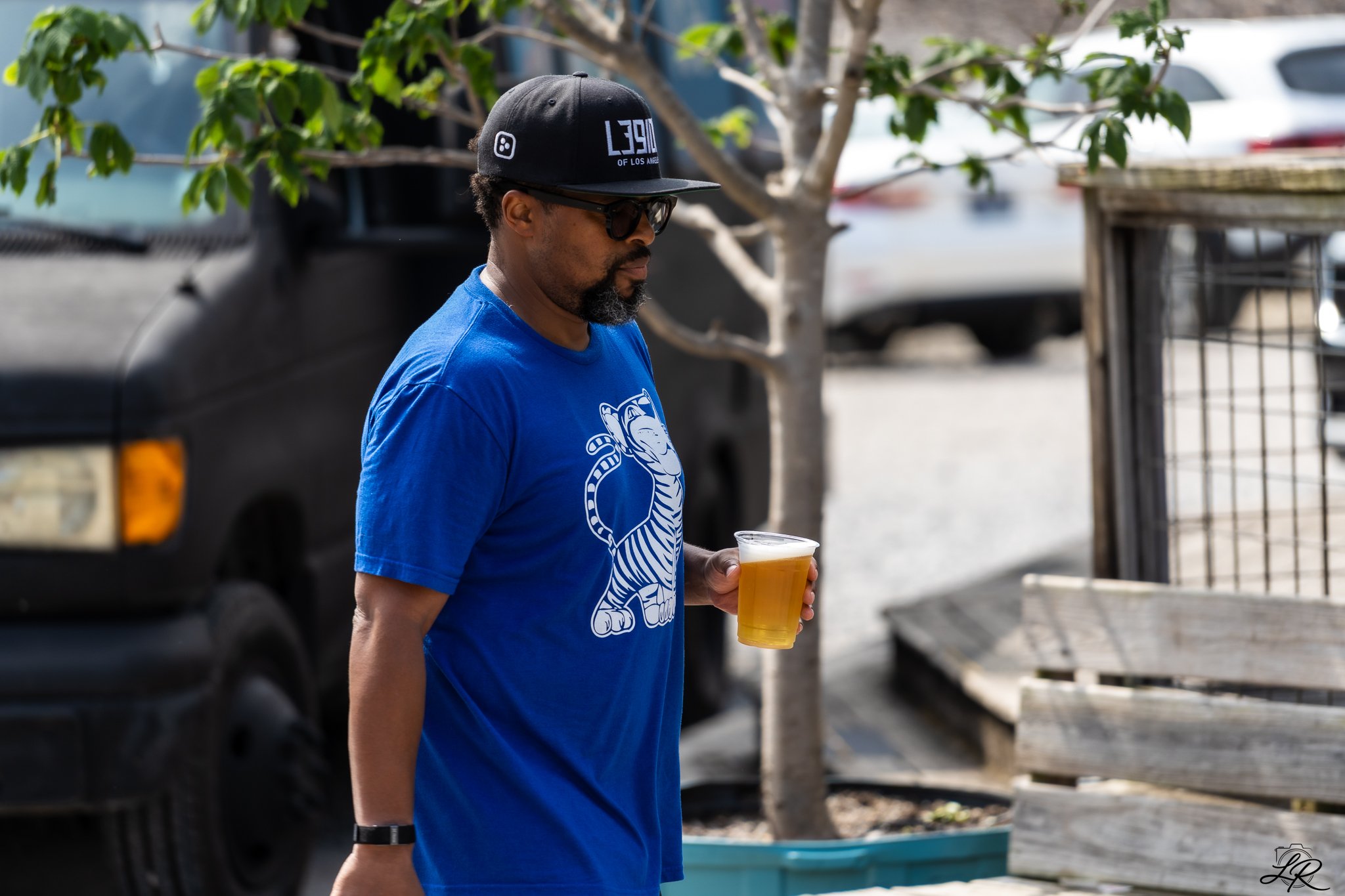 A man wearing a black baseball cap, sunglasses, and a blue T-shirt holding a cup of beer, walking outdoors near a tree and parked cars.