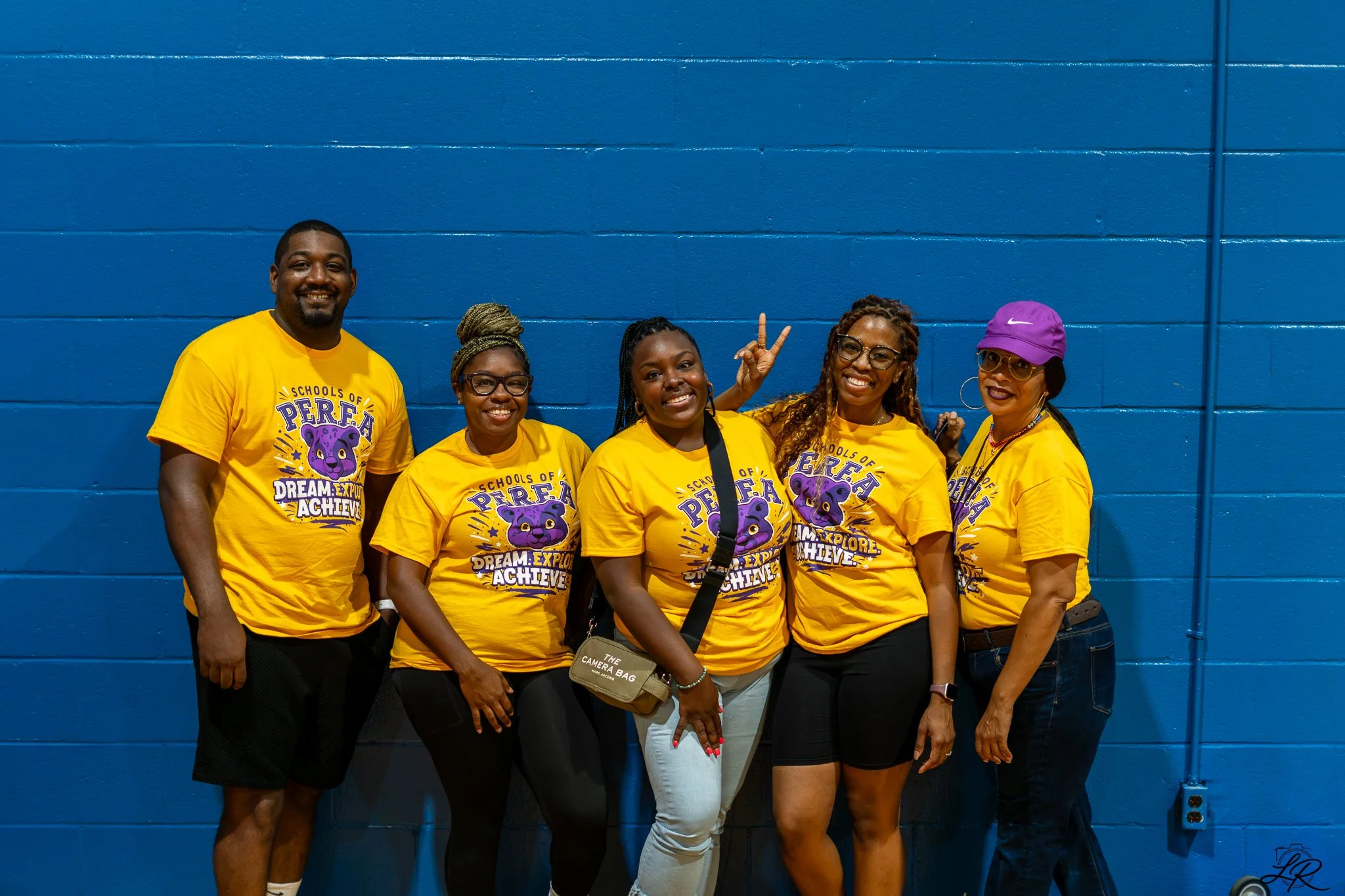 Six people standing in front of a blue cinder block wall, smiling, wearing yellow T-shirts with purple text and graphics for a school event, making peace signs and posing for a group photo.