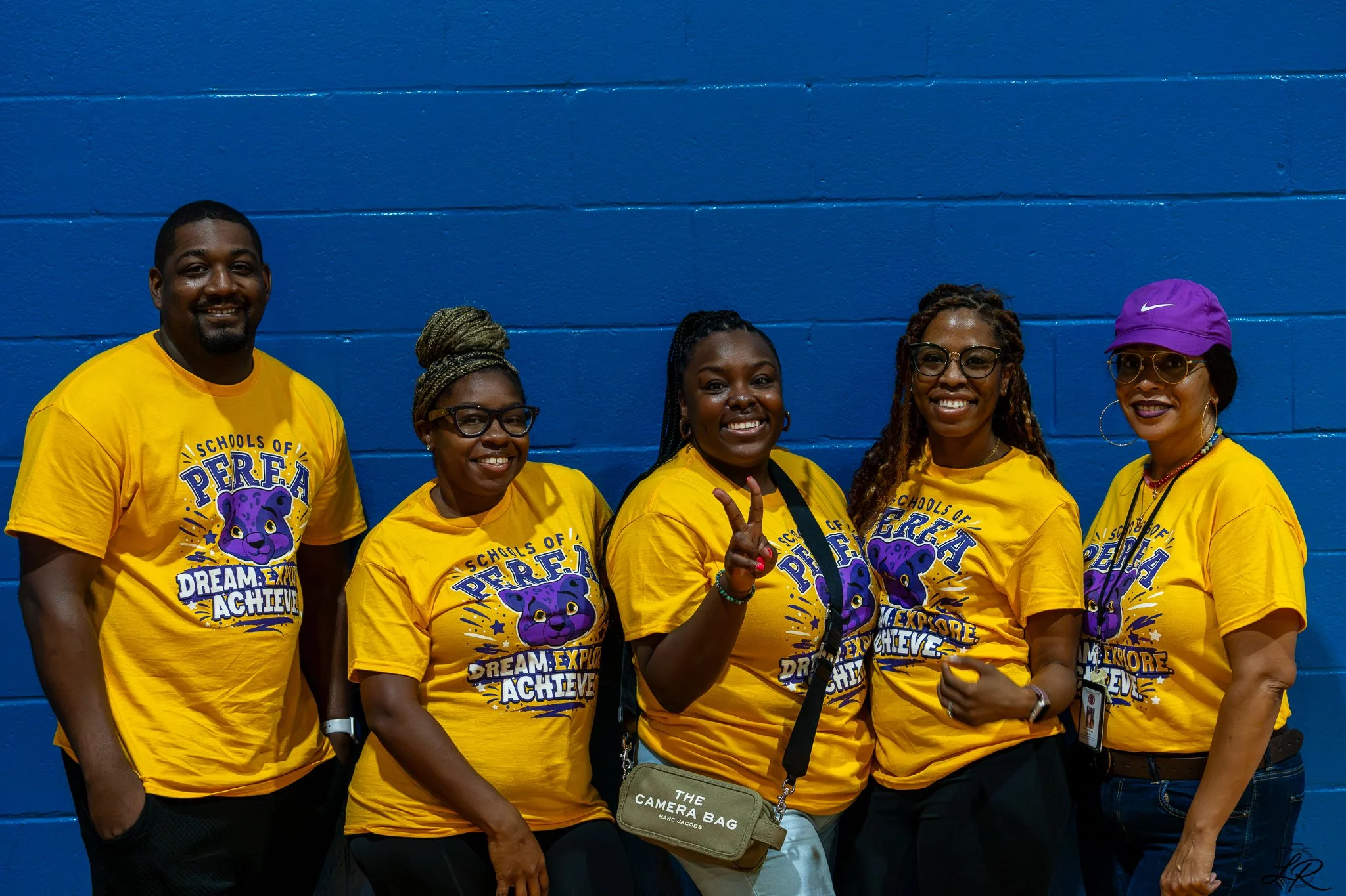 Group of six people standing in front of a blue wall, wearing matching yellow t-shirts with a purple panther logo and the text "Dream. Explore. Achieve." They are smiling and posing for the photo, with some making gestures and one wearing a purple Ni