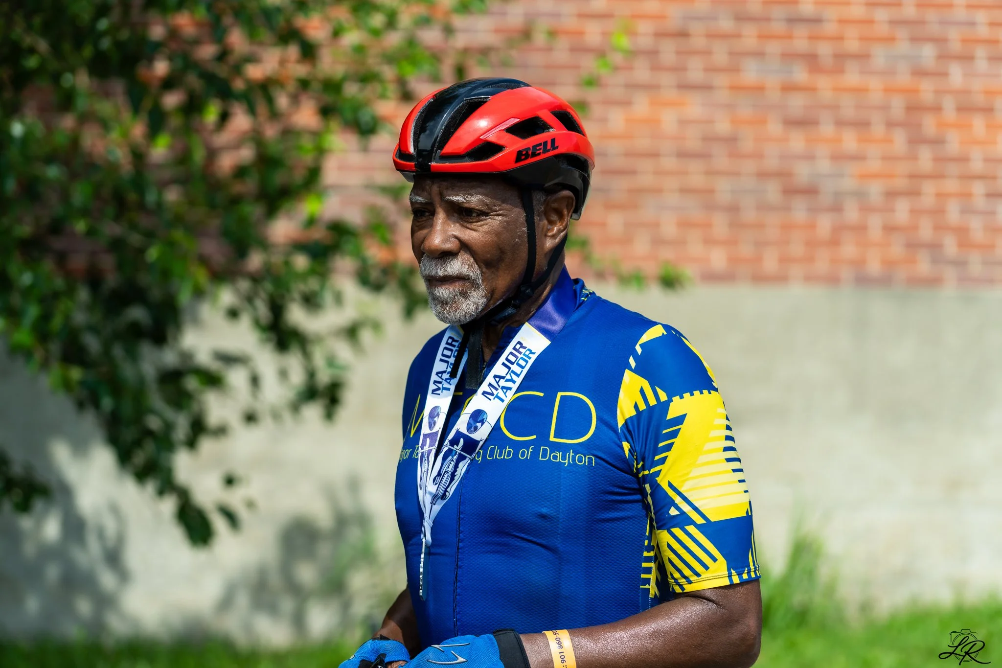 An elderly man wearing a red and black Bell cycling helmet and a blue racing jersey with yellow accents, standing outdoors against a brick wall and greenery.